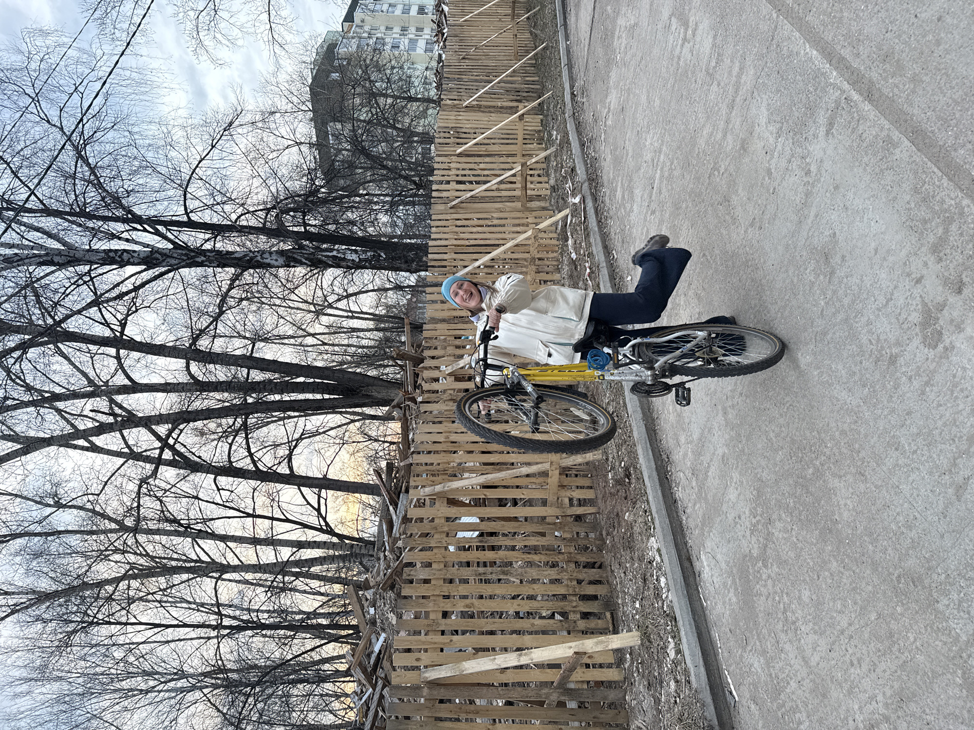 A young woman is photographed while riding a bicycle on a concrete path next to a wooden fence. The woman is wearing a blue beanie, a light-colored jacket, and dark pants. She is smiling at the camera. The bicycle has a yellow frame. Behind the fence is a line of bare trees and a multistory building, suggesting an urban or suburban setting in Chaikovsky, Russia, although specific identifiers are not evident to definitively confirm this location. The trees suggest it is late fall or winter. The ground is edged with dry grass and some debris.
