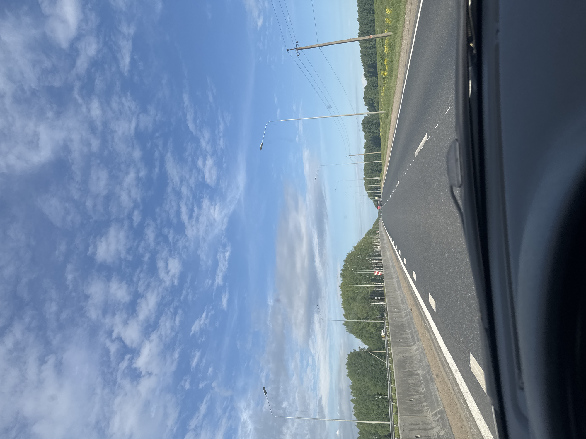 The image presents a daytime view from inside a moving vehicle traveling on a multi-lane highway, presumably in Selishcha, Russia. The bright blue sky dominates the upper portion of the frame, dotted with scattered, wispy white clouds, indicating clear and sunny weather.
The asphalt road, visible in the lower section, is marked with clear white lines, including a solid outer line and dashed inner lane dividers. A grey concrete or metal guardrail runs along the left side of the road. Parallel to the highway, a dense line of green trees, likely a forest or managed treeline, borders the landscape. Beyond these trees, a vibrant yellow field stretches towards the horizon.
Numerous utility poles with power lines and modern streetlights on tall, curved poles extend along the roadside into the distance. In the far distance on the highway, a small red car is visible, and behind it, possibly another light-colored vehicle, suggesting light traffic. A red and white striped warning sign, typical for road hazards, is faintly discernible on the left side of the road further down, though any text on it is illegible. The right edge of the image shows a dark section of the car's interior, from which the photo was captured. The overall scene depicts a peaceful and open road journey through a rural or semi-rural environment.