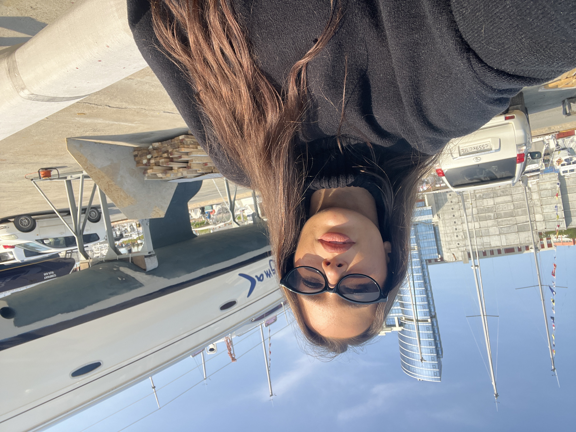 A woman with long, brown hair and sunglasses is taking a selfie in Vladivostok, Russia. She is wearing a black turtleneck sweater. Behind her is a marina with several sailboats, a white car with license plate "C559KC 125", a modern glass-paneled building, a crane, and a clear blue sky. The photo is taken on a sunny day.
