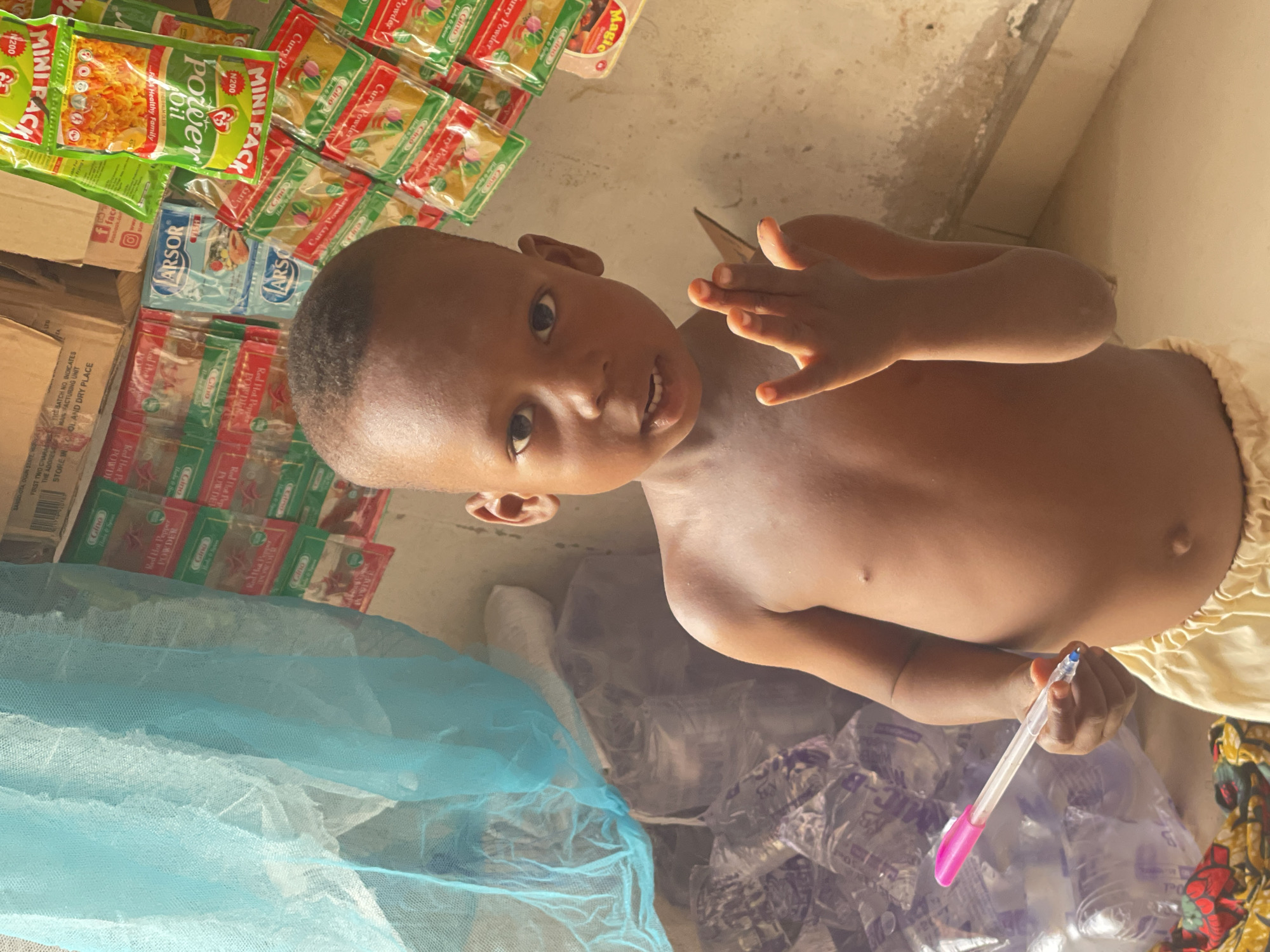 The image captures a slightly tilted, close-up perspective of a young child in Lokoja, Nigeria. The child, likely a boy with dark skin, short dark hair, and dark eyes, looks directly at the camera with a curious expression and a slight smile. He is shirtless, wearing light-colored lower garments, and holds a clear plastic pen with a pink cap in his right hand.
The background reveals an indoor setting, possibly a small shop or a home storage area, filled with various individually packaged food products. Prominently displayed are numerous "MINI PACK" sachets of "Power Oil Refined Palm Olein," some marked with "N200." Stacks of small pouches of "Gino" brand "Curry Powder" and "Red Hot Pepper Powder" are also visible, alongside "ARSOR" branded packets. Text such as "Magic," "Healthy Family," "STORE IN COOL AND DRY PLACE," and details about manufacturing are legible on the packaging and cardboard boxes.
On the lower left, a large piece of light blue netting or fabric is present, while crinkled clear plastic material is seen on the lower right, beneath the child's hand. The bright, even lighting suggests it is daytime. The scene portrays a casual moment, possibly within a local business or home environment, characterized by common household food items.