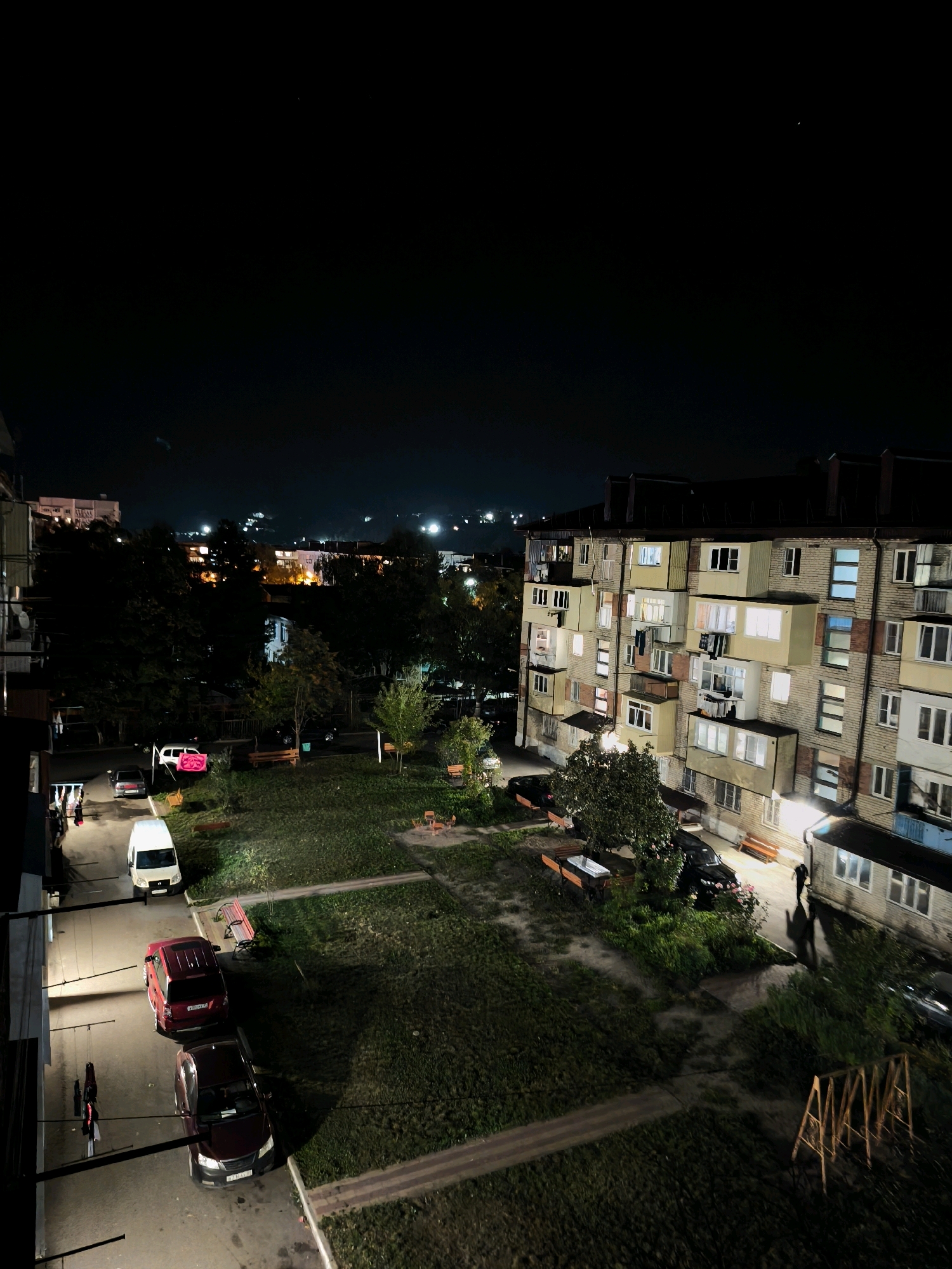 The image depicts a nighttime view of a residential courtyard in Karachayevsk, Russia. In the foreground, a paved area with parked cars and a white van is illuminated by artificial lighting. Beyond this, a grassy courtyard with several benches and a small children's play structure is visible. To the right, a multi-story apartment building with illuminated windows stands prominently, casting long shadows across the scene. In the distance, more buildings and streetlights suggest the presence of a town or city. The sky is dark, with a few faint lights visible on the horizon. A single person is silhouetted against the building on the right side of the image. The overall atmosphere is one of a quiet, urban night.
