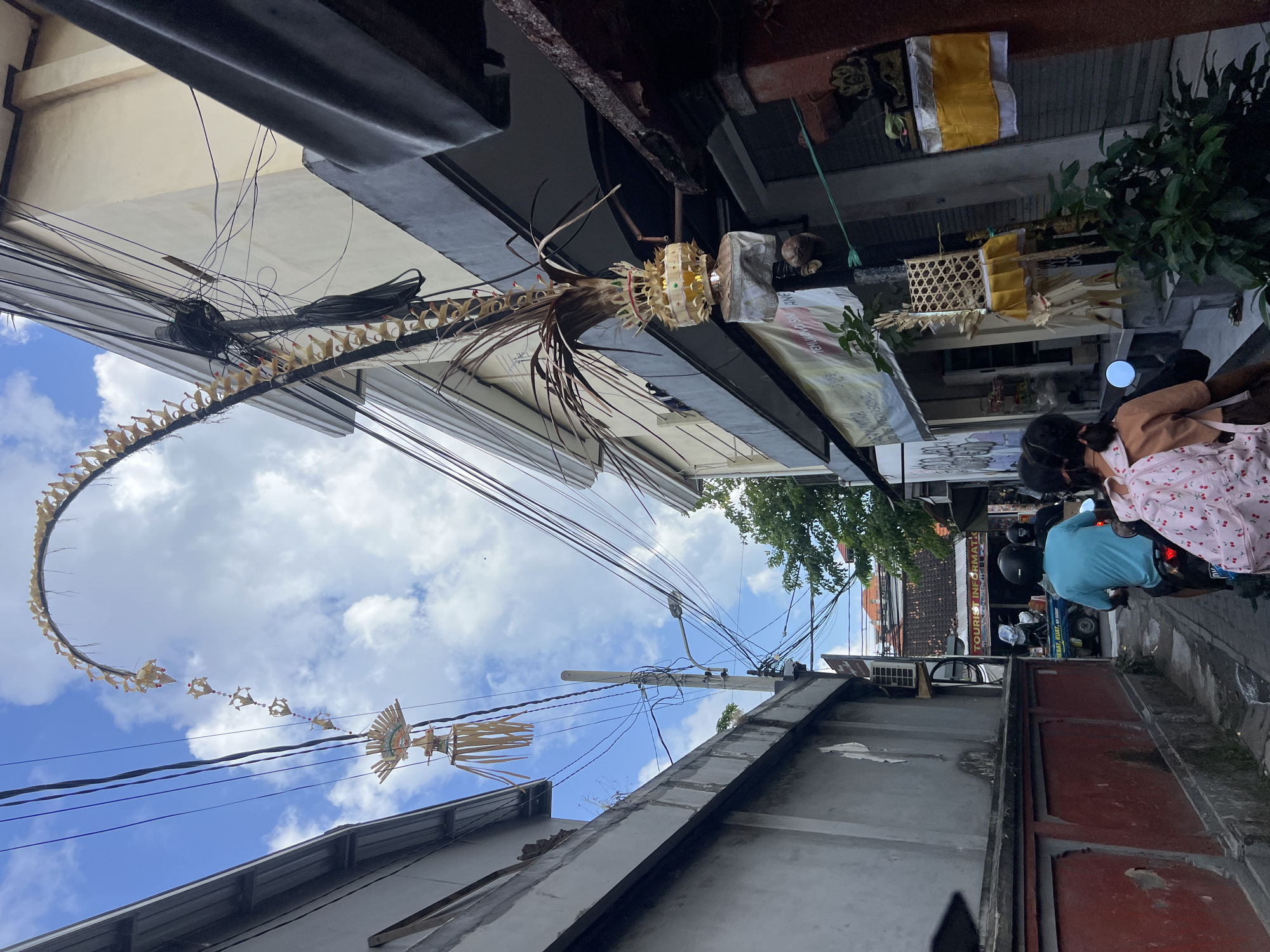 In Kuta, Indonesia, a narrow street is depicted with buildings on either side and a street lined with power lines. An ornate, woven structure, likely a Balinese Penjor decoration, arches high above the street, suspended by wires. Several smaller woven ornaments hang from the same wires. In the foreground, two people are riding on a scooter, facing away from the camera. The person in front wears a turquoise shirt and a helmet, while the person behind wears a pink backpack with cherry designs and a brown shirt. The scooter is moving down the street, which is paved and has a sidewalk. Shops line the right side of the street, with signs and awnings visible. The sky is blue with scattered clouds. The overall scene suggests a street in a populated area of Kuta.