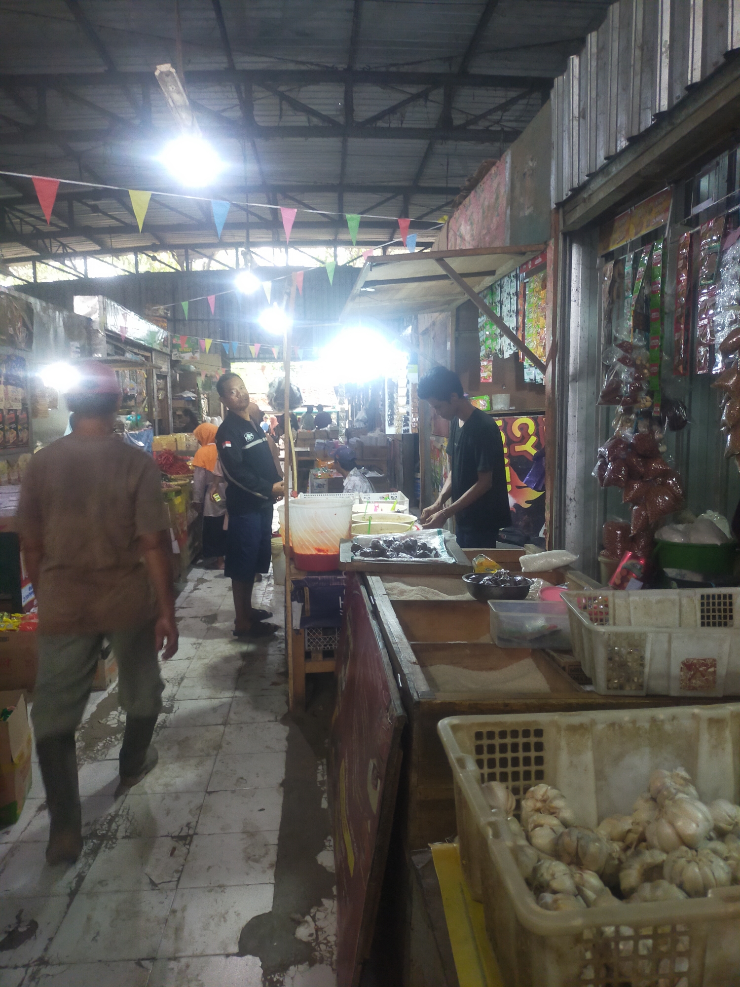 The image depicts an indoor market scene in Trayeman, Indonesia. The market appears to be a covered area with a corrugated metal roof supported by beams. Fluorescent lights are suspended from the ceiling, illuminating the space. Colorful triangular flags are strung across the market.
In the foreground, a person is walking away from the camera, wearing a brown t-shirt and grey pants, with dark boots. To the left, another person in a black jacket and blue shorts stands near a stall displaying various items, including what appears to be food products in containers. Further into the market, another individual in a black t-shirt is working behind a stall, arranging items on a surface. Stalls are lined with goods, including packaged food items, vegetables such as garlic in baskets, and other miscellaneous products. The floor of the market is tiled and appears wet in places, with visible puddles. The overall environment suggests a busy marketplace with active commerce.