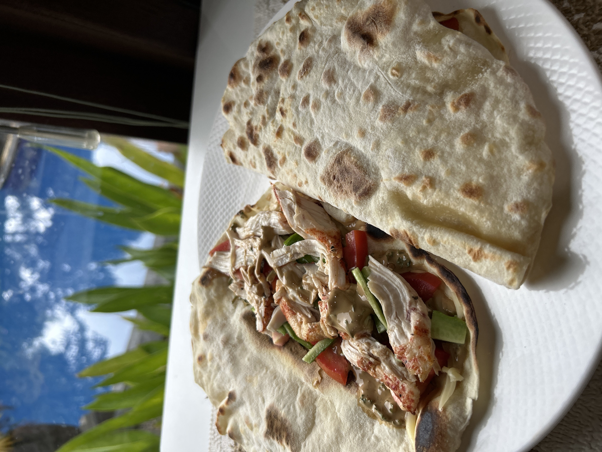 The image presents a close-up, elevated view of two filled flatbreads or wraps resting on a white, textured plate. The scene is indoors, with the plate positioned next to a window that reveals a blurred outdoor environment.
The main subject is the food. One flatbread is open, showcasing its vibrant filling. This includes shredded, light-colored meat, with some pieces exhibiting a reddish hue, suggestive of chicken, lobster, or crab. Mixed with the meat are diced red tomatoes and thin green slices, likely cucumber or green bell pepper. A creamy, light brown sauce generously coats the ingredients. The flatbreads themselves are pale with distinct, irregular brown spots from cooking, indicating they might be freshly made or pan-fried. The second flatbread is folded and placed beside the filled one.
The background through the window features a bright blue sky dotted with soft white clouds. Lush green foliage is visible at the bottom of the window view, suggesting an outdoor, natural setting. The natural light indicates it is daytime. Given the location context of Kuta Selatan, Indonesia, the scene likely depicts a meal in a tropical setting. There is no visible text in the image.