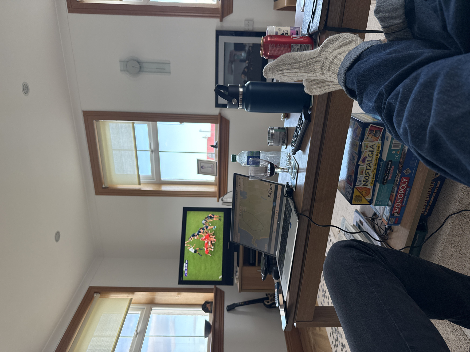 The image depicts a living room scene. In the foreground, a person is seated, wearing blue jeans and socks, with their legs extended onto a wooden coffee table. The coffee table is cluttered with various items including a laptop displaying a map with "5.43 km" visible, a blue water bottle, drinking glasses, board games ("Nostalgia," "Trivial Pursuit," and "Monopoly"), cans, and a remote control. In the background, a television displays a sporting event. Windows are visible in the background, revealing daylight. A guitar is on the floor near the table. The scene gives the impression of someone relaxing indoors.