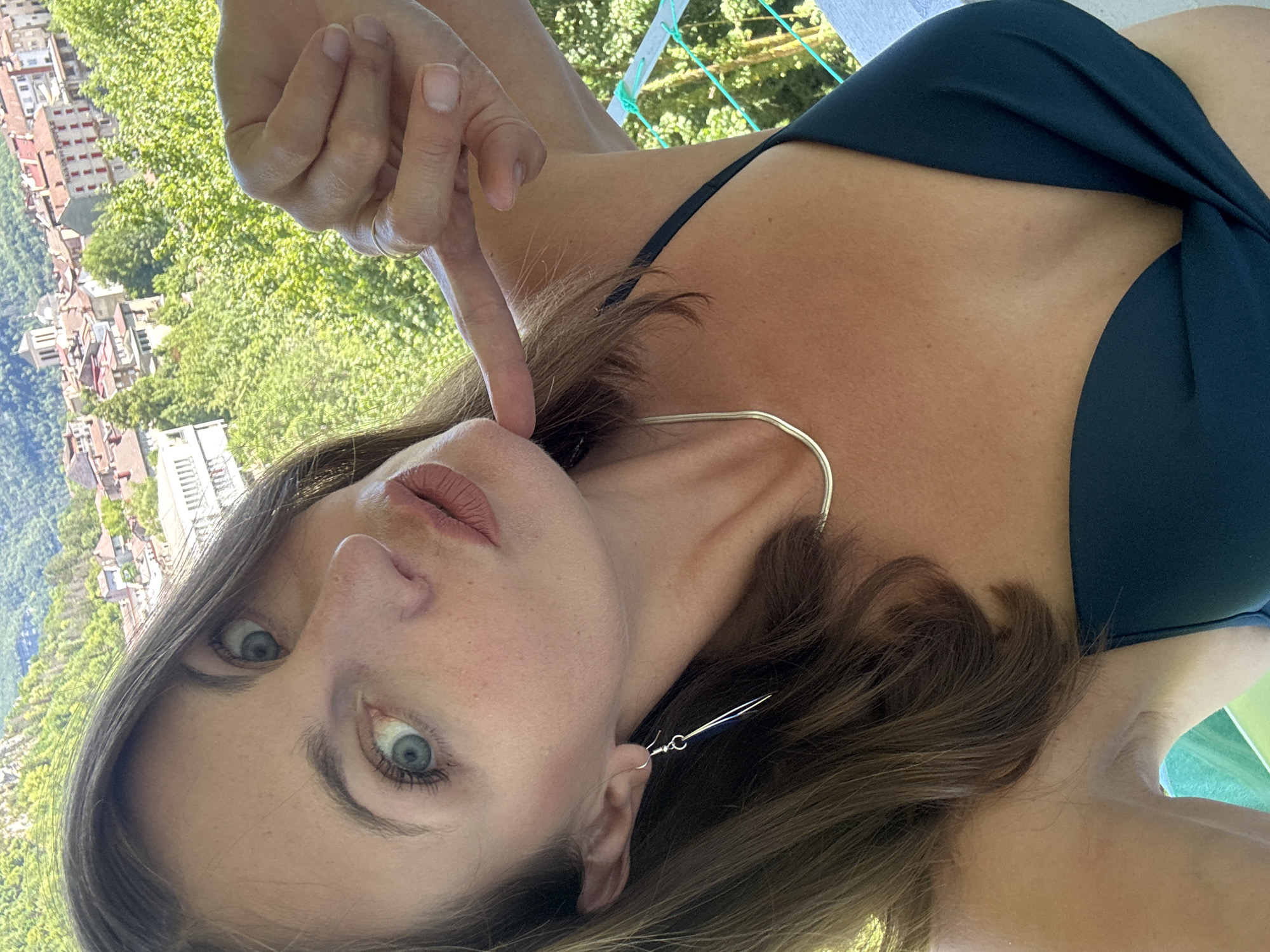 A woman with brown hair and blue eyes is taking a selfie with a city and forest in the background. She is wearing a dark green bikini top and a silver necklace. She has a silver earring in her left ear and is pointing at her chin with her right hand. The city appears to be Saint-Claude, France, with its characteristic red-tiled roofs. The background shows a lush green forest and mountains.
