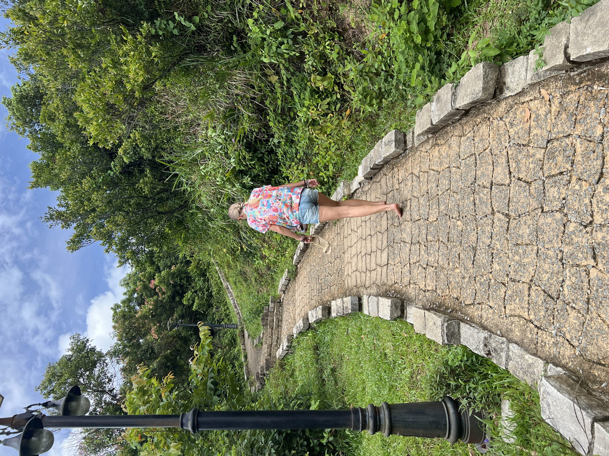In Đà Nẵng, Vietnam, a woman with blonde hair tied in a braid walks barefoot on a stone-paved pathway winding up a lush green hillside. She is wearing a colorful, floral-patterned short-sleeved shirt and denim shorts. She holds a pair of light-colored sandals in her right hand. The pathway is bordered by a low stone wall and surrounded by dense vegetation, including trees, bushes, and tall grasses. A vintage-style black lamppost stands to the left of the pathway in the foreground, and another lamppost is visible further up the path. The sky above is blue with scattered white clouds.