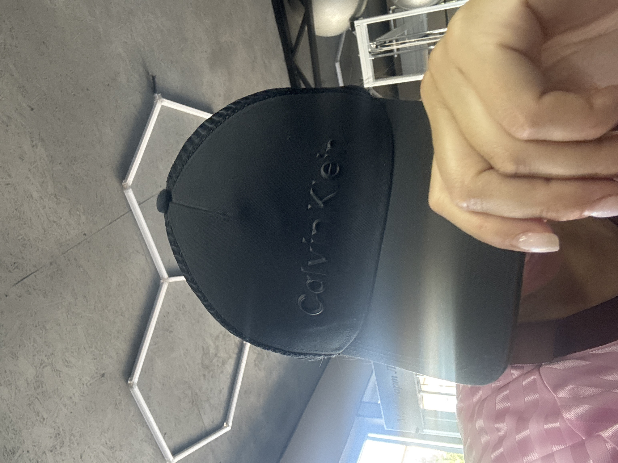A person is holding a black Calvin Klein baseball cap. The cap has a mesh back and the Calvin Klein logo embossed on the front. The person's hand, with manicured nails, is visible holding the cap. In the background, a hexagonal metallic structure is on a grey floor. Further back, there are industrial shelves with some round objects on them, and a glimpse of a light-colored ceiling. A pink patterned garment is visible in the lower right corner. The lighting suggests an indoor setting, possibly during the daytime. A sign with text is partially visible in the background.