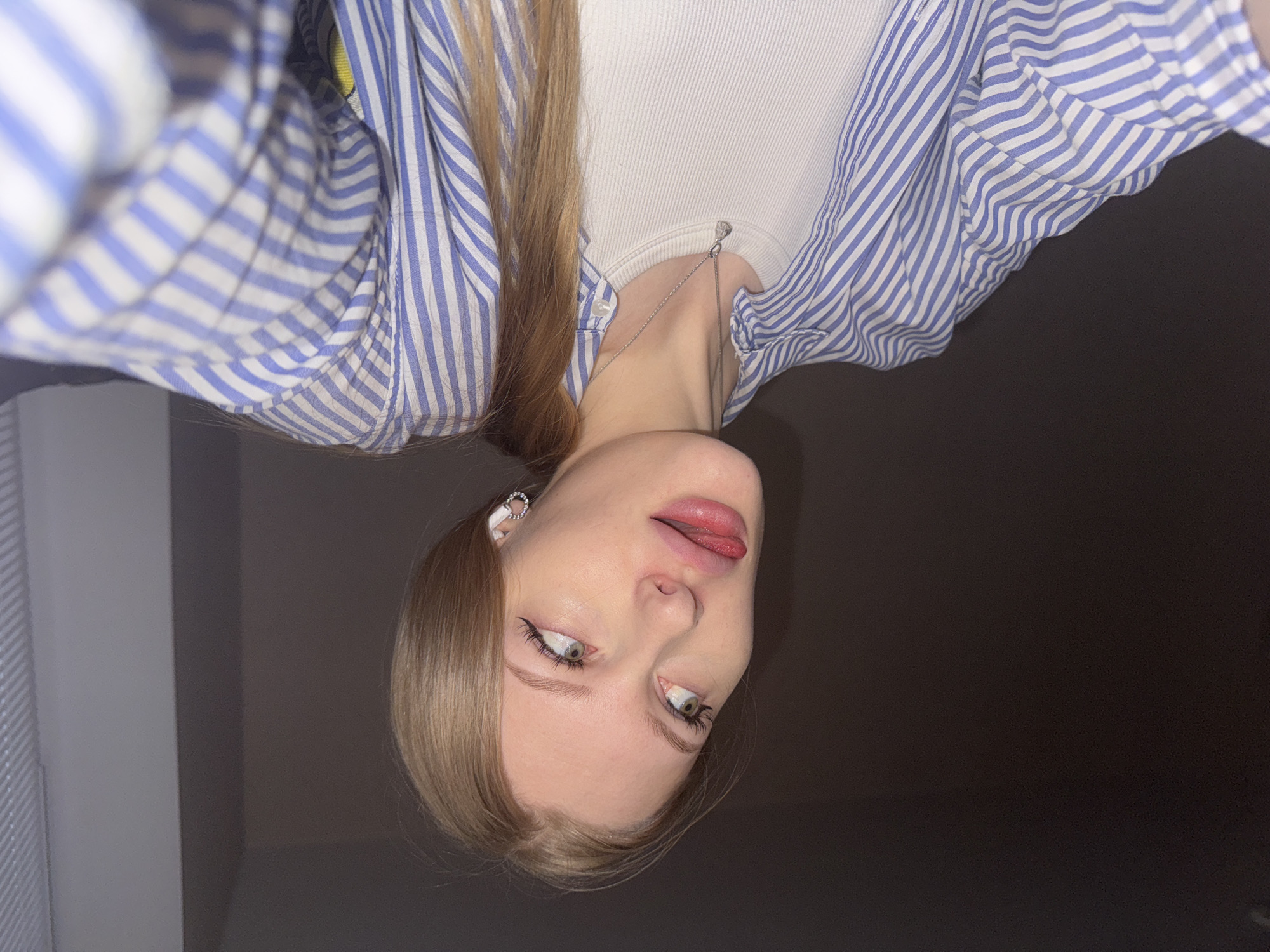A photograph shows a fair-skinned woman with light brown hair, wearing a white ribbed tank top and a blue and white striped button-down shirt. She is wearing a small silver necklace and a silver earring. Her eyes are green, and she has eyeliner. Her tongue is slightly visible between her lips. The photo is taken at an unusual angle, making the subject appear upside down. The background is dark gray. It is not possible to determine if the picture was taken in Perm, Russia from the image alone.