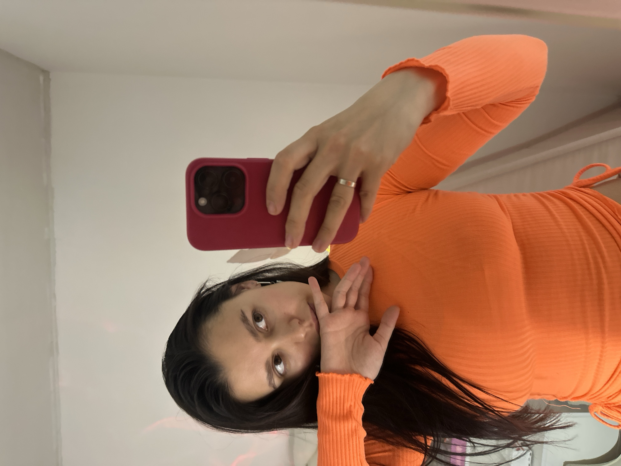 A young woman is depicted taking a mirror selfie indoors, in what is contextually specified as Chaikovsky, Russia. She has long, dark hair and is wearing a vibrant orange, ribbed long-sleeved top or dress that features ruffled cuffs. In her right hand, she holds a smartphone with a dark pink or red case, identifiable by its visible camera lenses, directed towards the mirror to capture the image. A simple silver band ring is worn on her right ring finger. Her left hand is subtly positioned near her face, with fingers lightly touching her chin and mouth area. She looks directly at the camera with a composed and neutral expression. The background is minimal, consisting of plain, light-colored walls, suggesting a simple room setting. The lighting is even and bright, consistent with an indoor environment that is well-lit. There is no visible text in the image. The provided media file is oriented horizontally, with the subject appearing on her side in the frame.