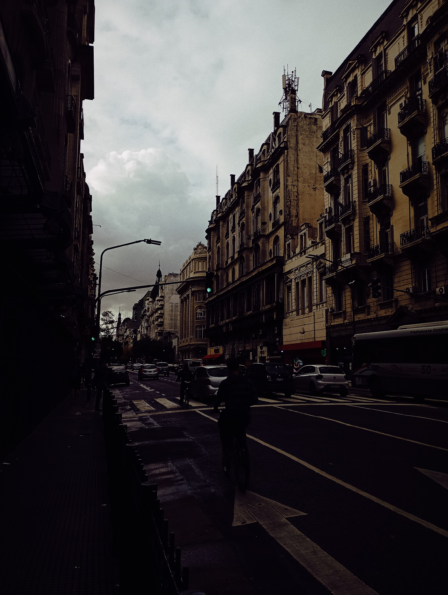 A low-light, atmospheric photograph captures a bustling urban street scene in Buenos Aires, Argentina, under a heavily overcast and gloomy sky. Tall, ornate buildings with classical architectural details, multiple stories, and decorative balconies line both sides of the street, giving it a European feel. A prominent cell tower or antenna array is visible on the rooftop of one of the buildings against the cloudy backdrop.
The street itself appears wet, reflecting the subdued light and indicating recent rain or damp conditions. Traffic includes several cars, some queued, and a large white bus partially visible on the right, bearing the number "50" and the text "Piedrabuena Congreso." A person on a bicycle is silhouetted in the foreground, riding towards the viewer, with a large white arrow painted on the asphalt indicating the direction of travel. Streetlights are present, with a green traffic signal visible in the middle distance. On one of the building facades, a red "ALQUILER" (For Rent) sign is discernible. The overall mood is dramatic and somewhat somber, emphasizing the city's architecture and the daily rhythm of urban life under a grey sky.