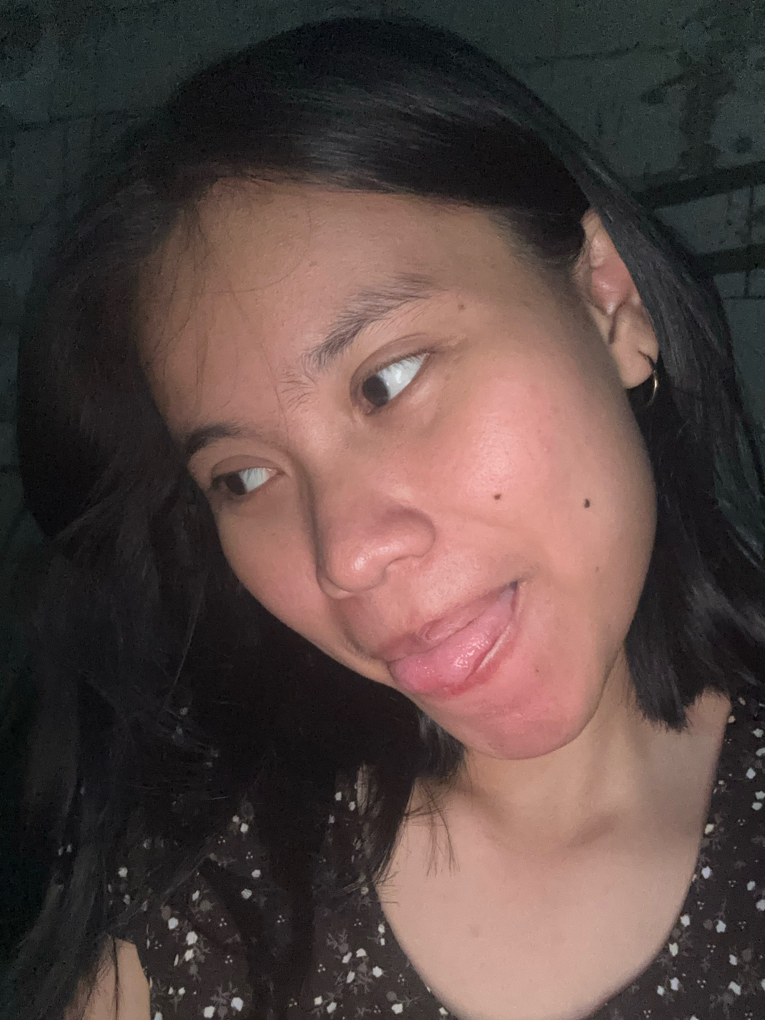 This is a close-up selfie of a young woman with dark hair and brown eyes, taken in low light. She is looking to her left with her tongue sticking out slightly. She is wearing a dark, patterned top. The background is dark and appears to be a textured wall. The location is not specified in the image.