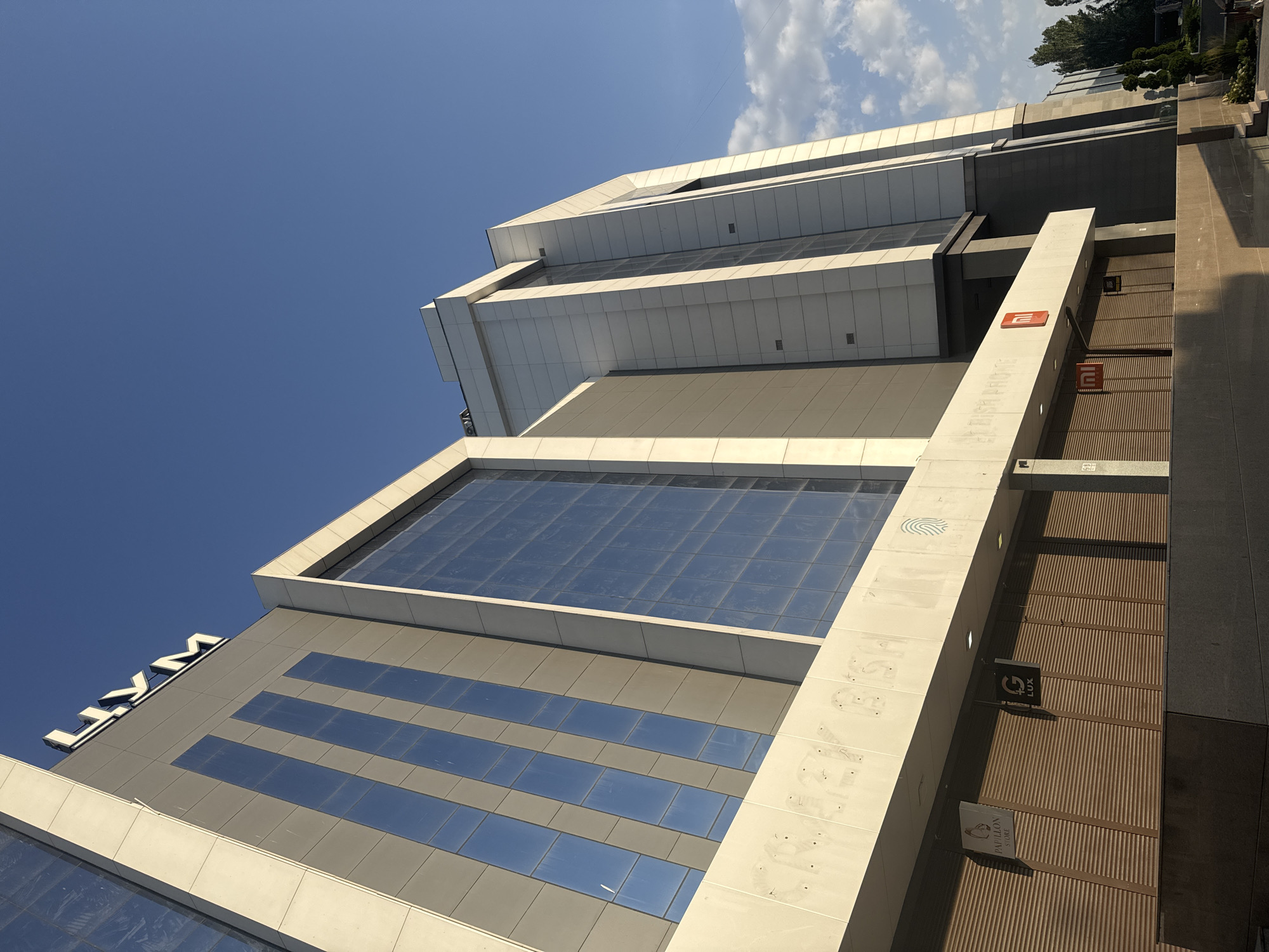 The image shows the facade of a modern, multi-story building in Bishkek, Kyrgyzstan, under a clear blue sky. The building features a combination of light-colored paneling and large, reflective glass windows that create vertical strips. The prominent signage at the top of the building spells out "ЦУМ" in large, block letters, indicating it is a department store. Below, the facade is punctuated by several closed storefronts with rolling metal shutters. Some of the storefronts have visible signage, including "PAPILLON STORE," "G LUX," and a red "mi" logo, suggesting these are individual retail establishments within the larger building. The lower part of the image shows a street-level view with the closed shutters, hinting that the shops are not currently open. The overall impression is of a large commercial building in an urban setting.