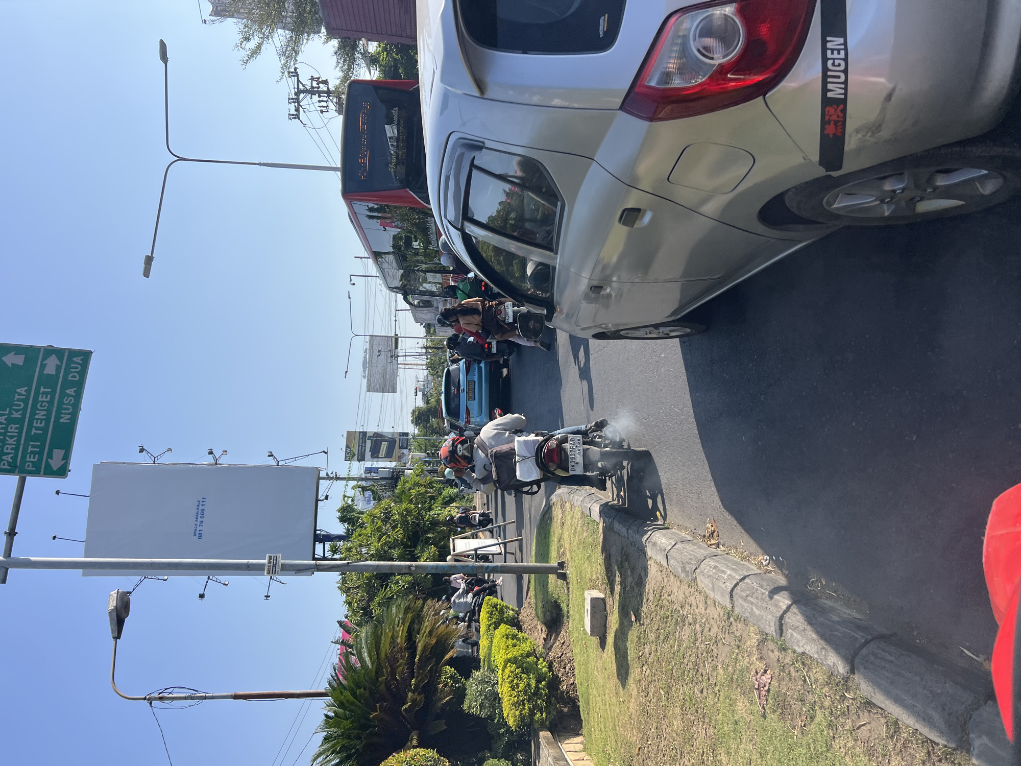 The image captures a street scene in Denpasar Barat, Indonesia, on a clear, sunny day. A silver car, possibly a Toyota Agya based on the sticker on the rear bumper that reads "MUGEN", is in the foreground, partially obscuring the view of the road ahead. The car has a sticker with Japanese characters above "MUGEN". Several motorcycles are visible in traffic, with one in the immediate foreground on the right side of the frame, emitting a cloud of exhaust. A person on this motorcycle is wearing a helmet and a backpack. Further up the road, other vehicles, including a bus and a light blue car, are also present in the traffic. The environment includes a green median with manicured bushes and trees, a sidewalk, and a concrete curb. Street signs are visible, including one directing traffic with text in Indonesian, indicating directions to "PARKIR KUTA" and "PETI TENGET" and "NUSA DUA". A large, white billboard with a phone number "081 70 668 111" is also prominently displayed. Power lines and streetlights are part of the urban infrastructure. The overall scene depicts a typical traffic situation in a busy urban area.