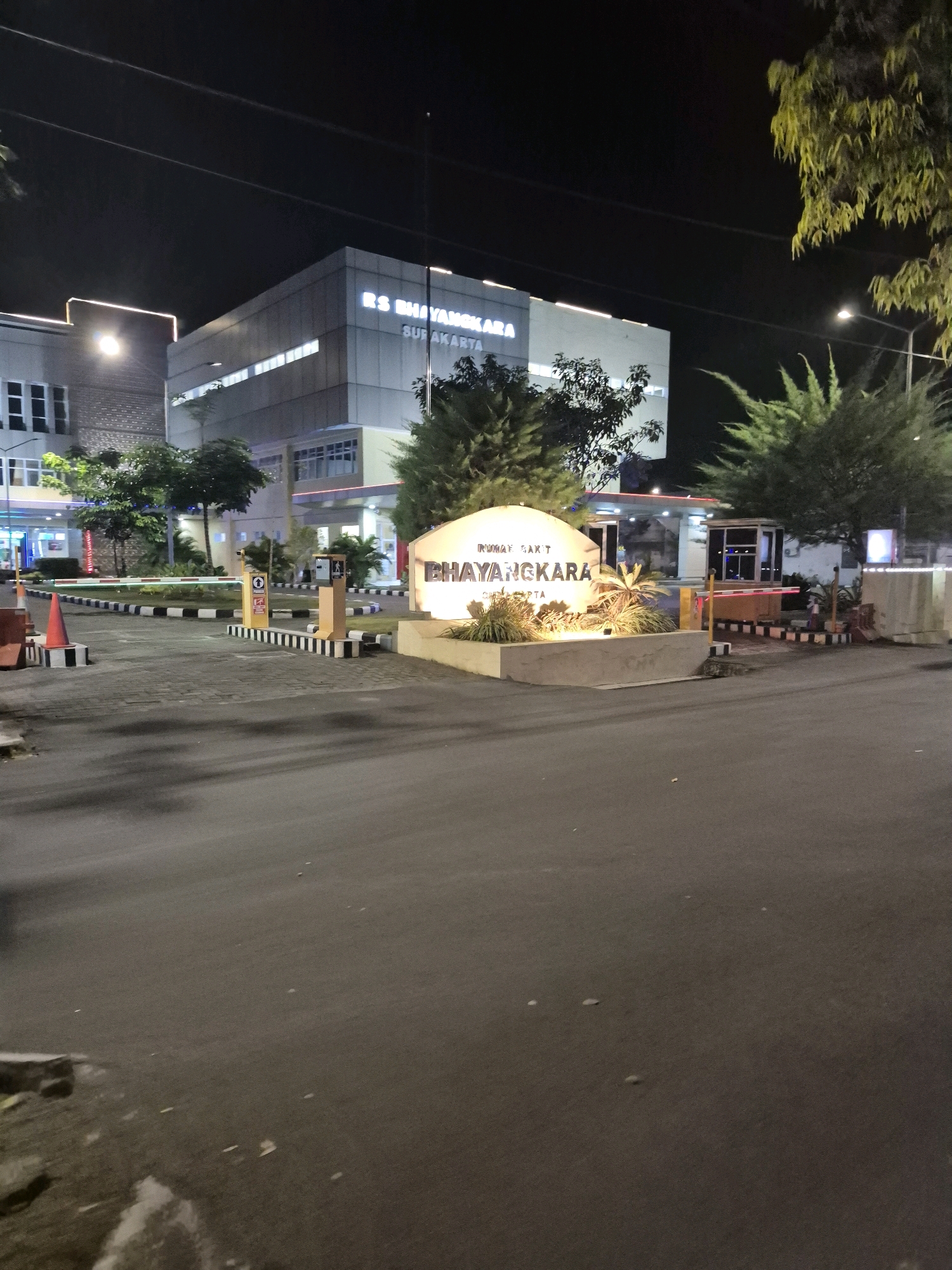 The image depicts the entrance to RS Bhayangkara Surakarta, a hospital located in Surakarta, Indonesia. It is nighttime, and the scene is illuminated by artificial lights. The hospital building is visible in the background, with its signage lit up. In the foreground, there is a road leading to the hospital entrance, with a barrier gate and a security booth. A landscaped area with a sign that reads "RUMAH SAKIT BHAYANGKARA SURAKARTA" is also prominent. The environment appears to be a paved road with some vegetation and trees. The overall scene is that of a well-lit hospital entrance at night.