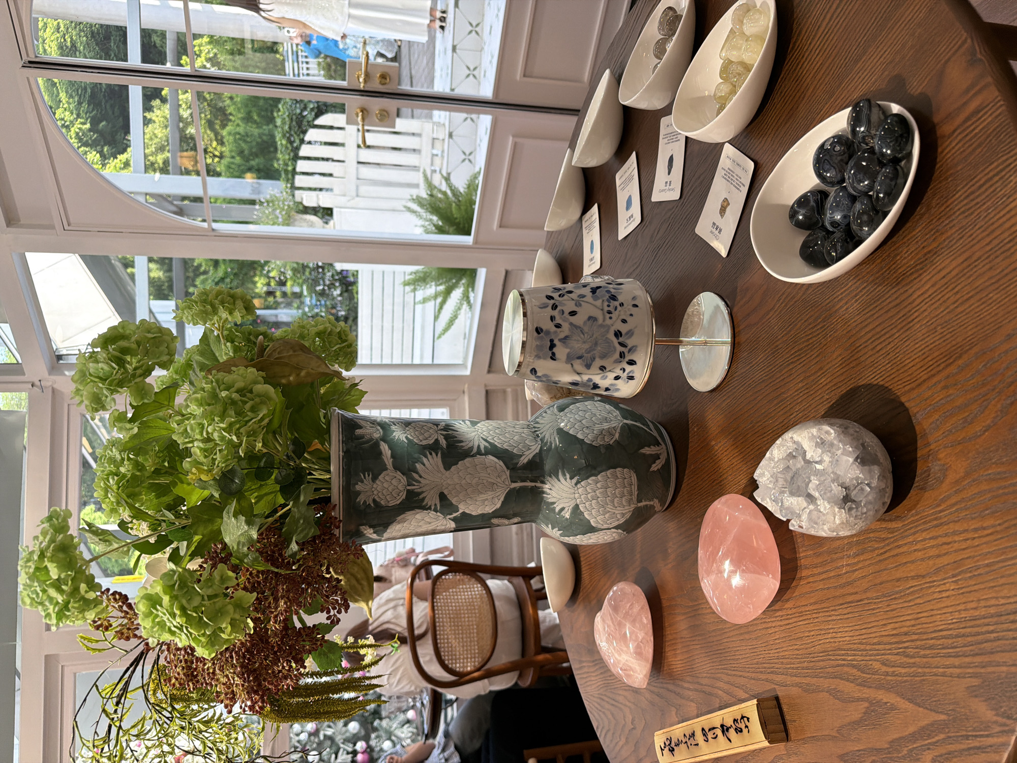 The image depicts a curated display on a dark wood table, possibly in a shop or studio, within Liangjiang, China. A vase of green hydrangeas and brown foliage dominates the lower left, with a pineapple-patterned vase. To the right of the vase are various objects arranged on the table: a blue-and-white patterned cup, a silver candleholder, several pink rose quartz stones, a geode, several white ceramic bowls containing stones (one contains black stones), and small name cards with Chinese characters. The table is situated near a window with a white frame, offering a view of a garden with a white picket fence. In the background, a person is partially visible, wearing a white dress. The environment appears well-lit and upscale.
