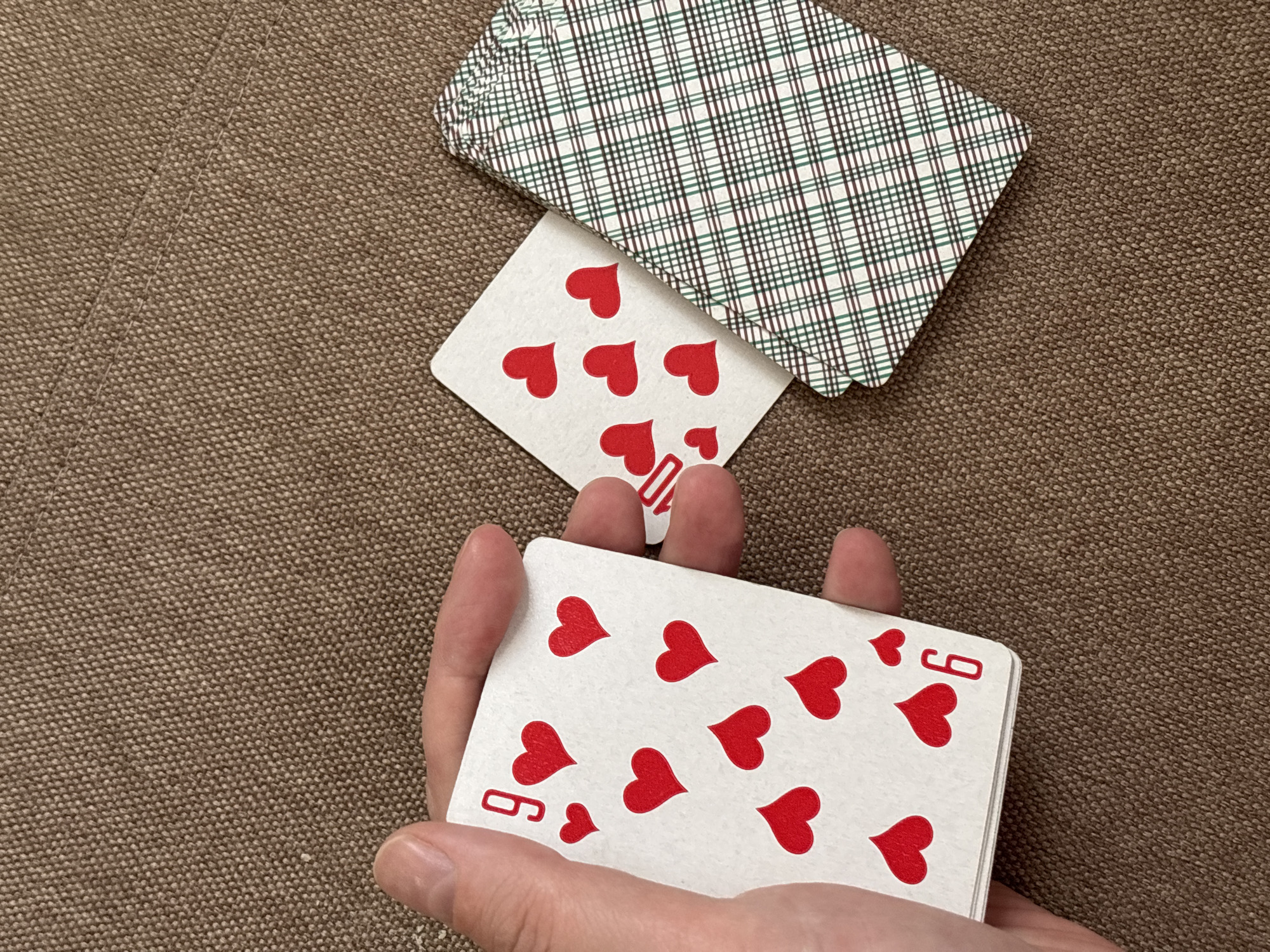 The image shows a close-up of a hand holding a stack of playing cards. The top card is the 9 of hearts. Next to it is a 10 of hearts. In the background is a deck of playing cards face down with a green and white plaid pattern. The surface is a brown fabric. There is no discernible information in the image that suggests the location is Chaikovsky, Russia.