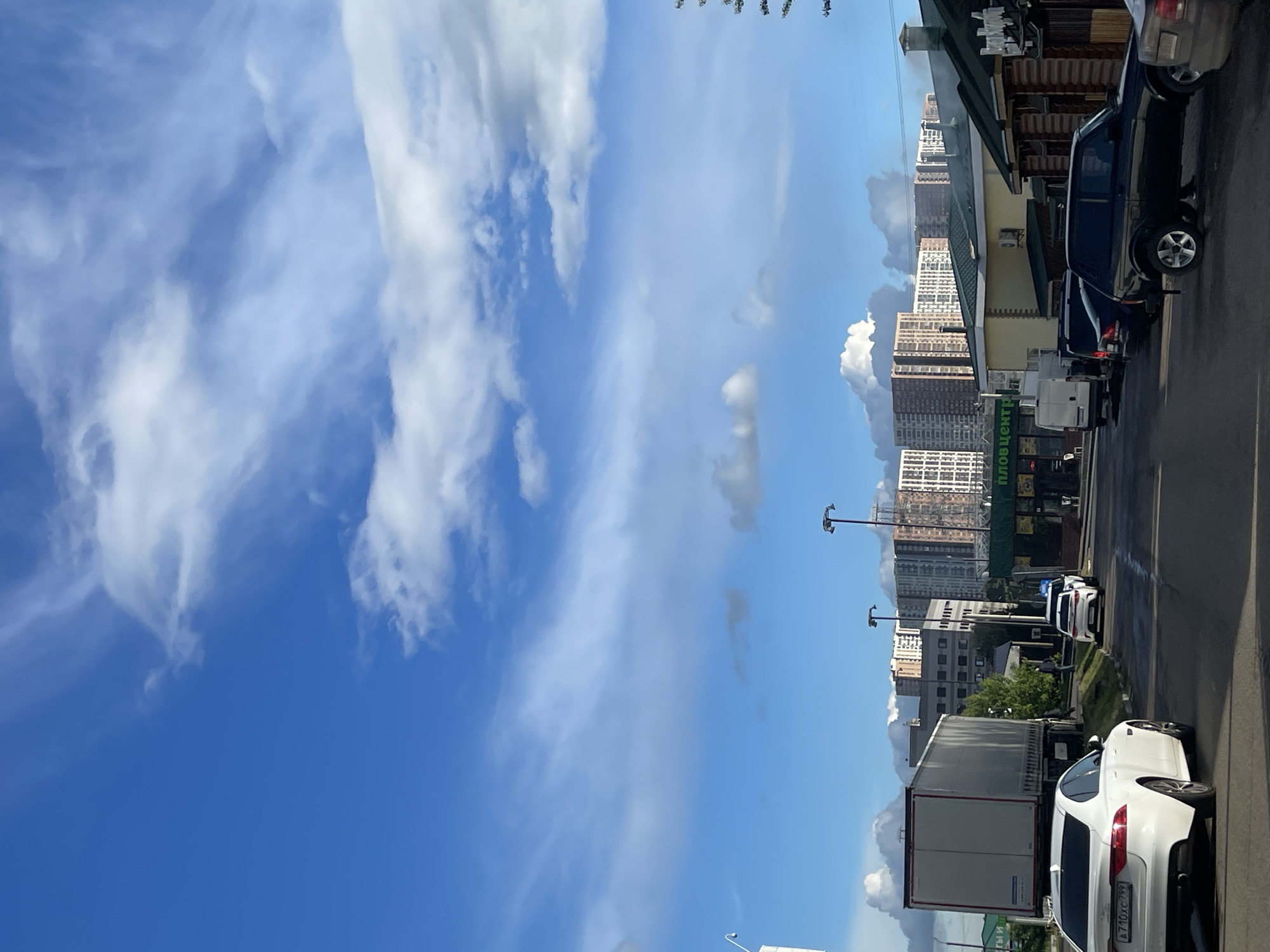 The image captures an urban scene in Bittsa, Russia. A clear blue sky with wispy clouds dominates the left side of the frame. On the right, buildings and various vehicles line a street. These vehicles include cars and a truck. The buildings appear to be a mix of residential and commercial structures. Street lights are also visible along the road.