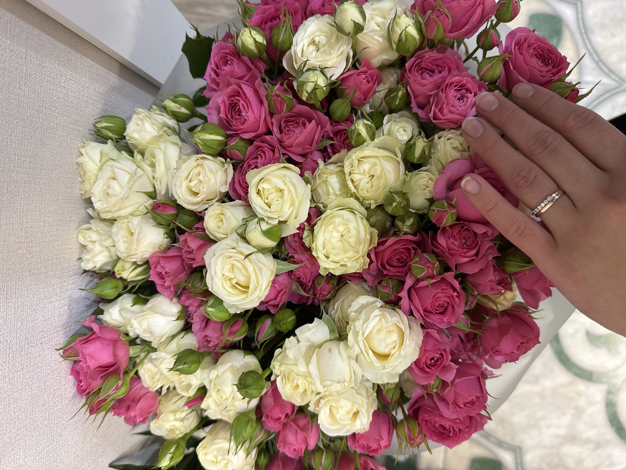 The image displays a close-up of a vibrant bouquet of roses, primarily featuring a mix of white/cream and fuchsia/deep pink colored blooms. Numerous green buds are also visible among the open flowers, indicating various stages of growth. The roses are densely packed, forming a lush arrangement.
On the right side of the bouquet, a person's hand, likely female, is gently placed on or near the flowers. The hand has natural, unpolished nails. A double-banded ring is worn on the fourth finger of the left hand; one band appears to be plain metal, and the other is adorned with a line of small, clear stones.
The setting is indoors. To the left, a textured wall or panel with a light gray or beige hue provides a backdrop. To the right and bottom, a lighter surface with a subtle, geometric pattern in light green or gray is visible, suggesting a floor or tabletop.
The scene captures a moment of appreciation or presentation of the floral arrangement. No specific time of day or weather is discernible. There is no visible text in the image. The context of the image is Kazan, Russia.