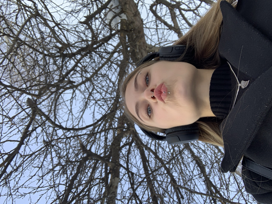 An upward-angle selfie captures a young adult female from the chest up, positioned outdoors in Chaikovsky, Russia. The individual has long, light-colored hair and light blue eyes, with her lips puckered into a "kiss" pose. She wears black over-ear headphones, a black turtleneck sweater, and a black overcoat. Prominent facial piercings include a silver septum ring, a silver labret piercing centered on her lower lip, and a silver horizontal eyebrow piercing above her right eye. A silver necklace with a stylized pendant rests on her turtleneck. The background consists of numerous bare tree branches silhouetted against a clear blue sky, indicating a late autumn, winter, or early spring setting.