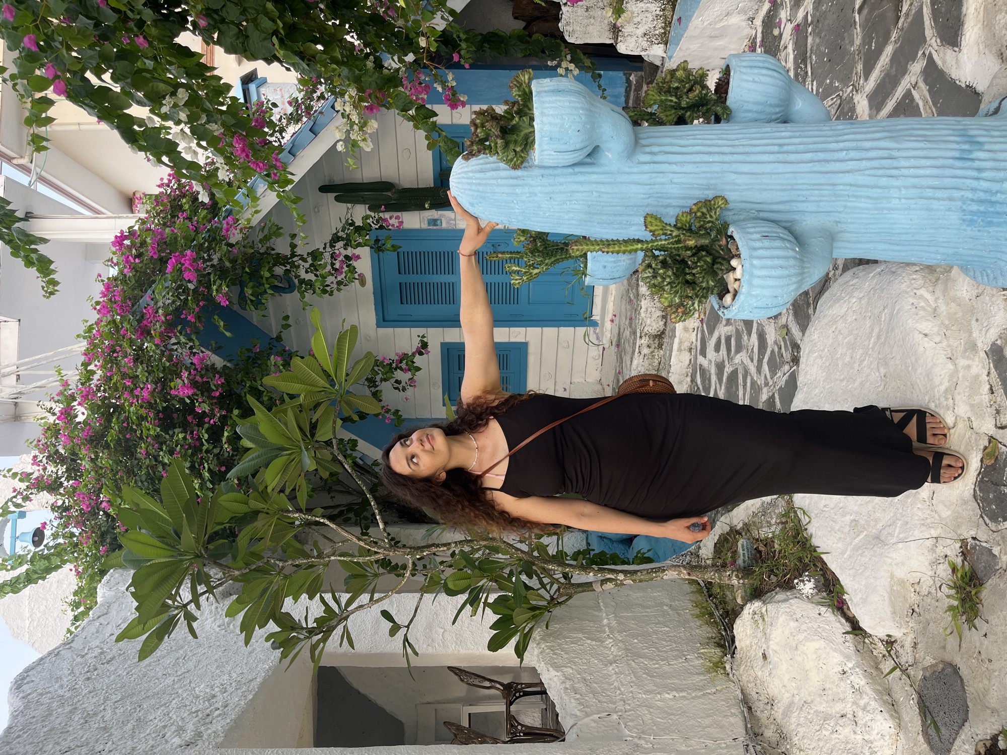 A woman in a black dress stands outdoors, her arm extended and touching a light blue, cactus-shaped planter. She has long, dark, curly hair and is looking upwards with a slight smile. She is wearing a necklace and a brown cross-body bag. The planter is in front of a white building with blue shutters and decorative elements. Pink and white flowers cascade from an overhead structure. The ground is a mosaic of white and dark stones. The scene appears to be in a garden or courtyard. The lighting suggests it is daytime, and the weather seems pleasant. There is no visible text.