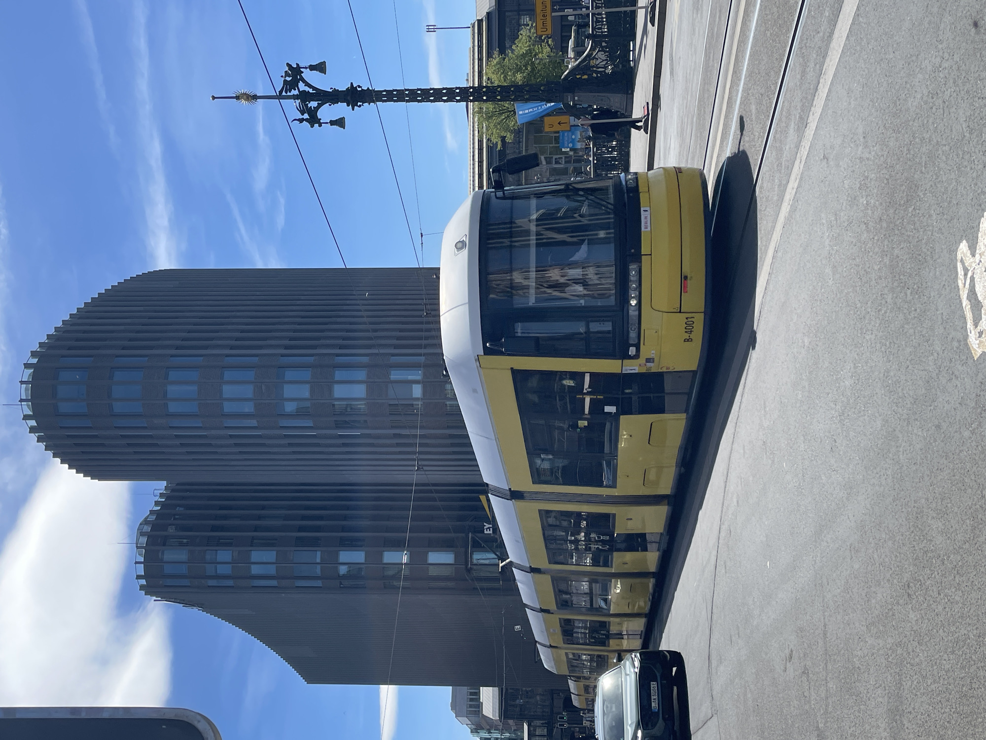 The image displays a bright, sunny urban scene in Berlin, Germany. A modern yellow and white tram, characteristic of the city's public transport, is prominently featured, traveling along its tracks. The tram carries the identification "B-4001" on its side and "BERLIN" vertically near its front.
In the background, against a clear blue sky with a few scattered clouds, stand two distinctive, dark grey modern buildings. These high-rise structures feature curved, ribbed facades and numerous rectangular windows. A decorative black street lamp post, adorned with bell-like features and a sun emblem at its peak, stretches across the upper right.
Overhead lines for the tram are visible, as are the tram tracks on the asphalt road. On the sidewalk to the right, a person is seen in the distance, and a yellow sign with the German word "Umlenkung" (diversion) and an arrow is discernible, indicating road changes. A small part of a black car is visible at the bottom right, with a partially obscured license plate. Strong shadows on the road suggest it is daytime, likely mid-morning or afternoon, under a clear sky. The scene depicts a typical bustling yet calm city environment.