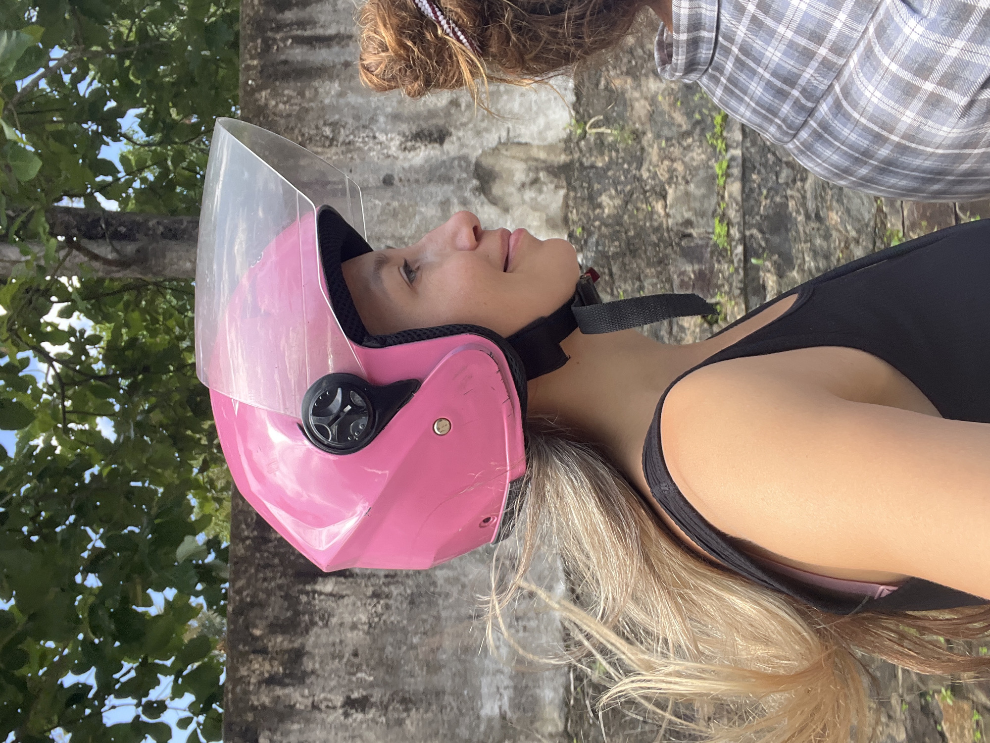 A woman wearing a pink helmet and a black tank top is riding on a motorcycle. She is looking to the right and smiling. Her hair is blonde and long. Another person is visible on the right, wearing a plaid shirt. They are riding on a street in Kabalana, Sri Lanka, with a stone wall and trees in the background.