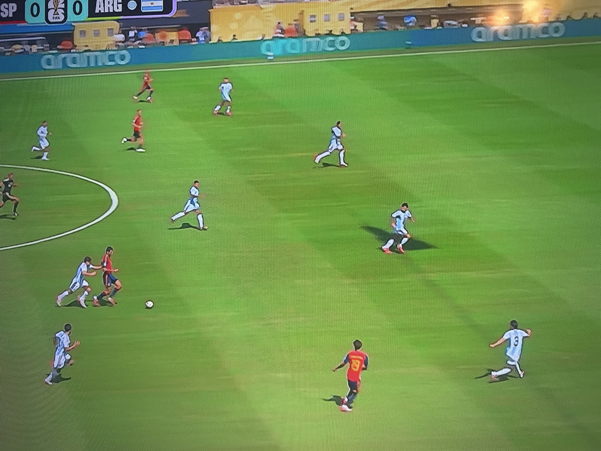 The image shows a soccer match in progress on a bright green field. The score displayed at the top of the screen indicates "SP 0" and "ARG 0". There are several players on the field wearing white and light blue uniforms, and some players in red uniforms. The players are actively engaged in the game, with some running, some kicking the ball, and some in defensive positions. The environment appears to be a professional sports stadium, with a light blue advertising board along the side of the field displaying the "aramco" logo. In the background, there are spectators in the stands. The location in Lapės, Lithuania is not explicitly depicted or inferable from the image itself; it is information provided contextually.