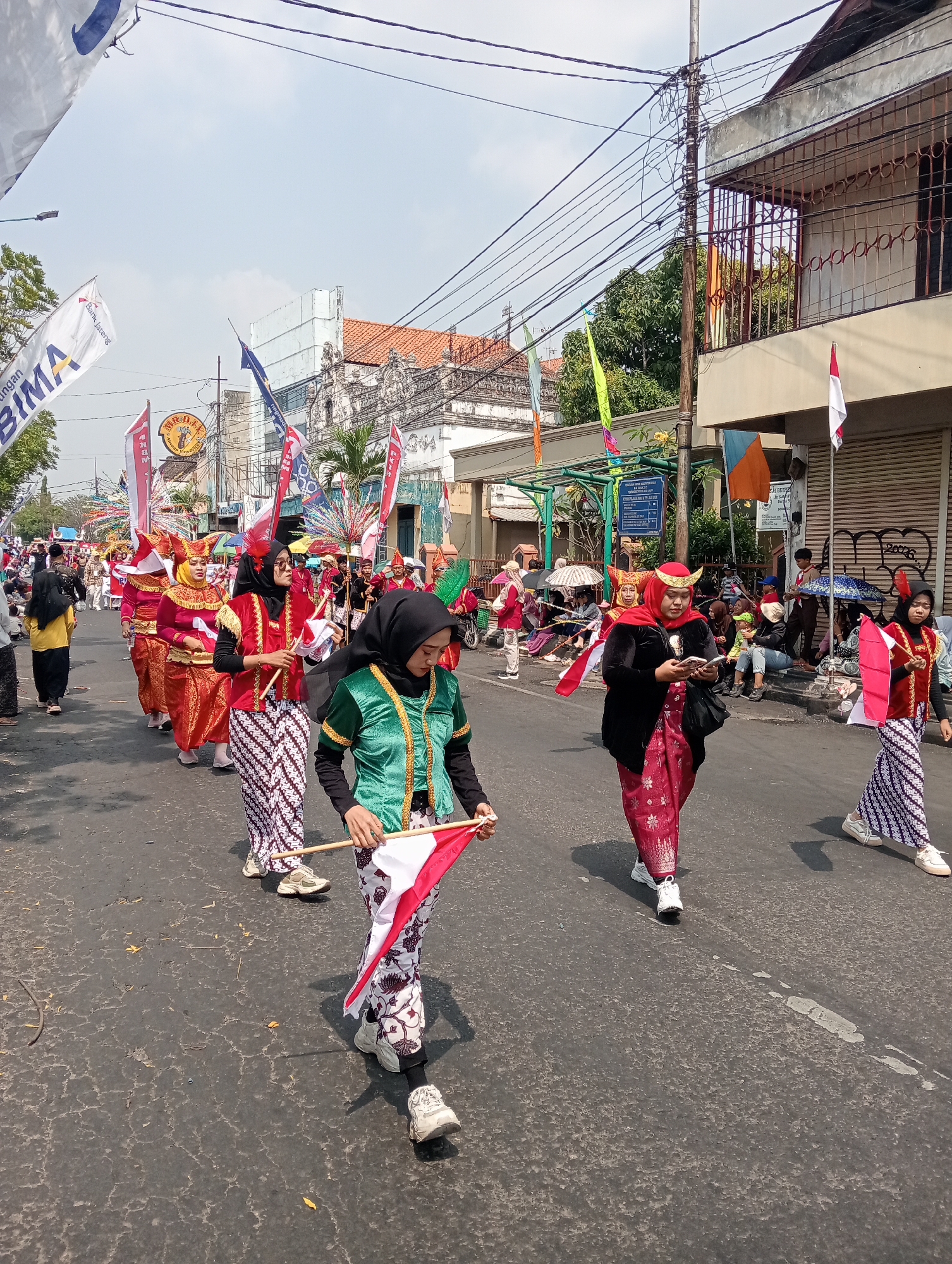 In Bintoro, Indonesia, a procession is underway on a street, characterized by participants in traditional attire. The foreground features individuals adorned in vibrant red and green outfits, some carrying small Indonesian flags. The women wear batik-patterned sarongs and headscarves, with some accessorizing with decorative headdresses. Behind them, more participants continue the parade, accompanied by onlookers seated on the side of the road and others standing further back. The street is lined with buildings, some exhibiting ornate architectural details, and is decorated with various flags and banners, suggesting a festive or ceremonial event. Overhead, power lines crisscross the sky, and trees are visible in the background. The weather appears to be sunny.