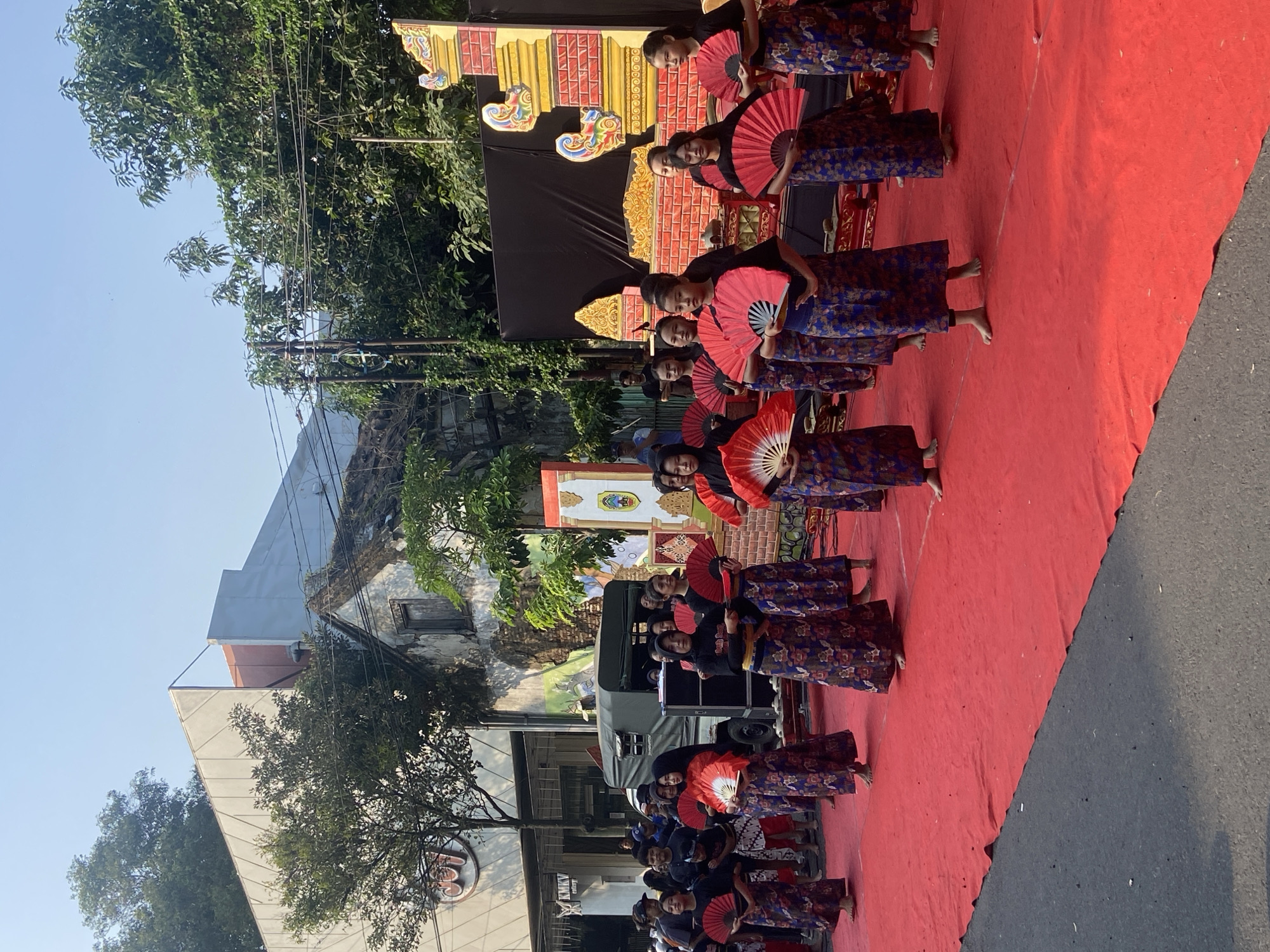 This is a medium shot of a group of young women in traditional Indonesian attire performing a dance on a red carpeted stage outdoors. The women are wearing dark tops and patterned sarongs in blue and red. They are all holding open red fans, which they appear to be using as props in their dance. The stage is set up in front of a backdrop that includes a black curtain and a decorative, brightly colored structure with yellow and red accents. In the background, there is a street with some parked vehicles and buildings, including a modern building with a large sign that reads "KMKV ENTERTAINMENT." There are also trees and power lines visible, indicating an urban or semi-urban environment. The sky is clear and blue, suggesting it is daytime. The overall impression is of a cultural performance or festival taking place in a public space.