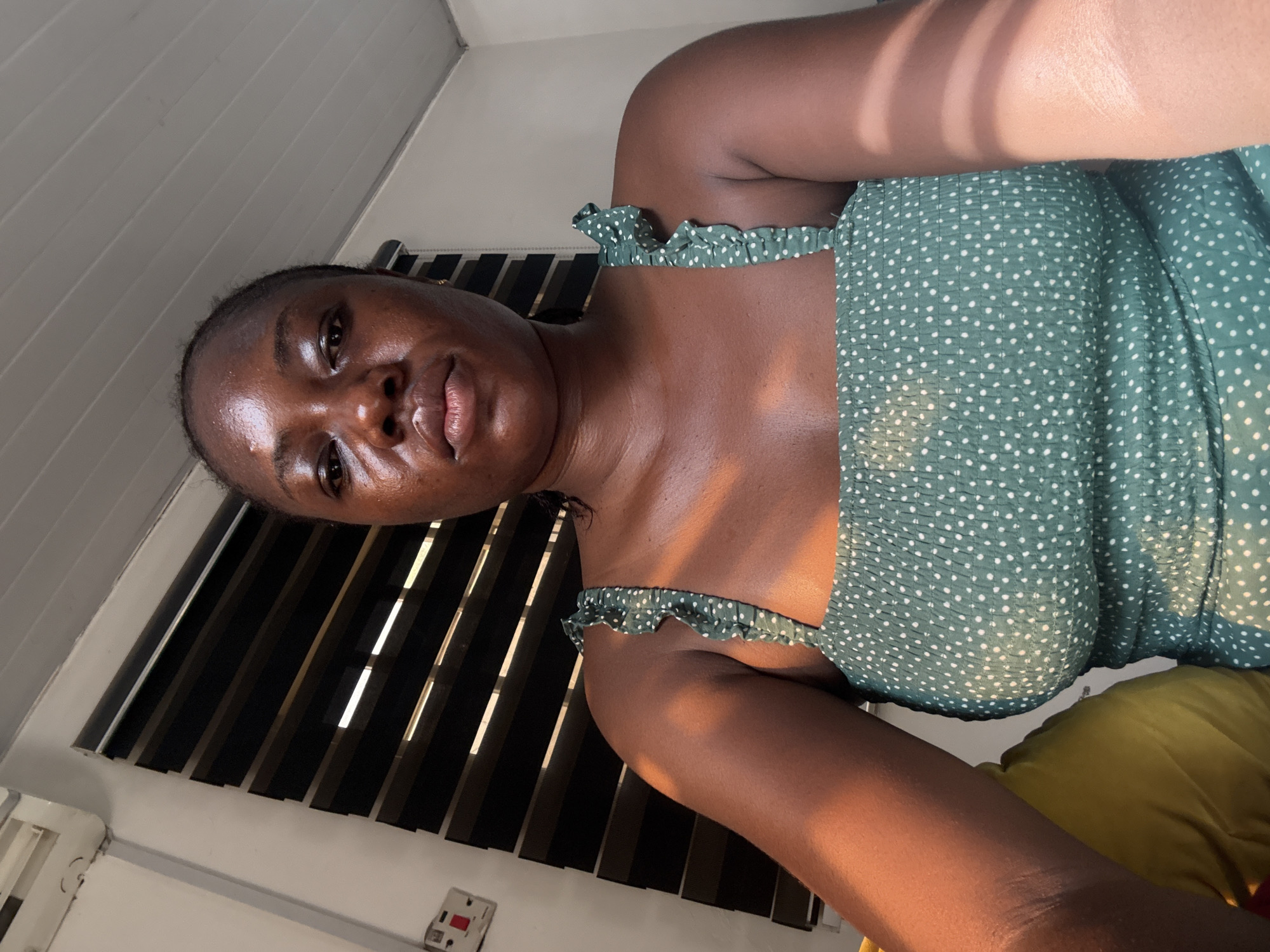 A medium shot depicts a dark-skinned woman wearing a green polka-dot top. The background includes a wall with Venetian blinds. The location is likely indoors, possibly in Abuja, Nigeria, but specific details to confirm the exact location are absent.