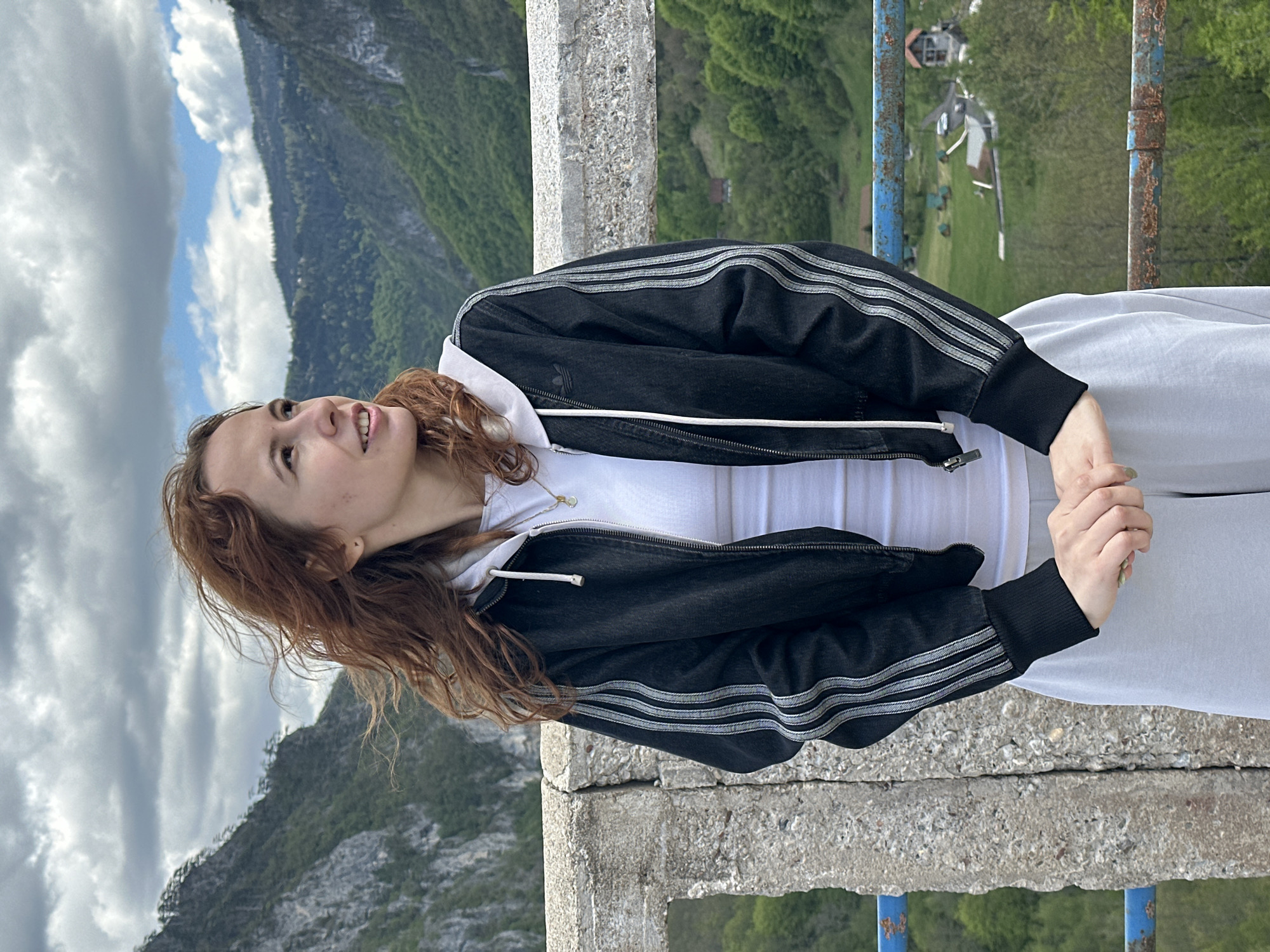 A young woman with long, reddish-brown hair is standing on a concrete structure in what appears to be a mountainous area. She is wearing a white hooded sweatshirt and a black jacket with white stripes on the sleeves, and gray pants. She is looking upwards and to the left. The background features a mountainous landscape with a green valley and some buildings. In the distance, a partly cloudy sky is visible. The blue railing is present on the bridge or platform she is standing on. Based on the appearance of the mountainous background and buildings, it is possible that the scene is located in the vicinity of Đurđevića Tara, Montenegro.