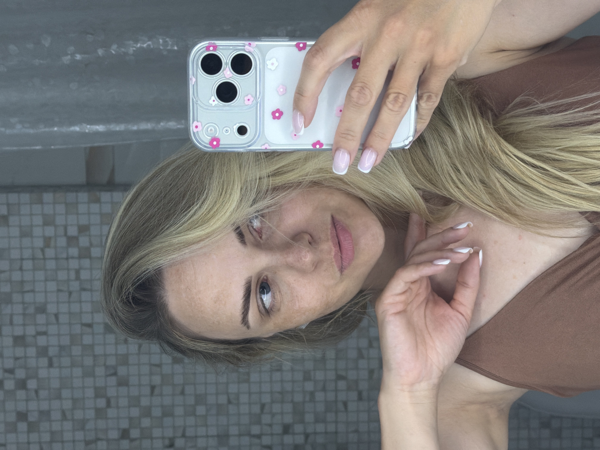A woman is taking a selfie in a bathroom with a grey-tiled floor and a white sink or counter visible in the background. She holds an iPhone in her left hand, which is decorated with a clear case featuring pink and white flowers. Her nails on both hands are painted with a French manicure. She has fair skin, blonde hair, and freckles. Her gaze is directed toward the camera. Her top is brown.
There is no information in the image to determine that this image was taken in Votkinsk, Russia.