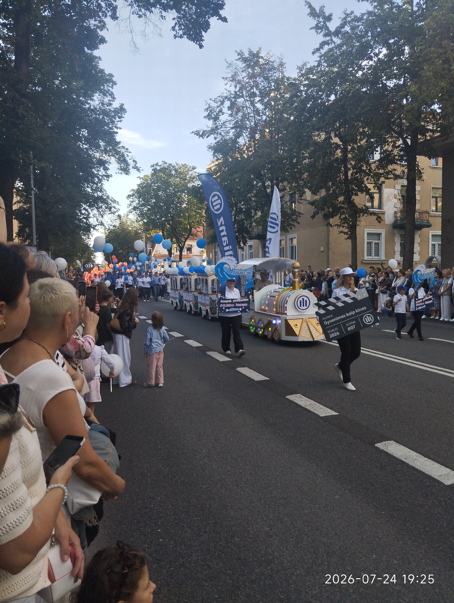 The image captures a street parade in Klaipėda, Lithuania. A decorated miniature train, likely a float, is moving down the asphalt road. Several people are walking alongside the train, some holding signs. One woman in the foreground is holding a clapperboard that reads "Gyvenimas kaip kinas" (Life as cinema), "Dublis: 2026", and "Scena: #07". In the background, a crowd of spectators lines the street, with trees and buildings forming the urban environment. Balloons, mostly blue and white, are visible throughout the scene. The timestamp "2026-07-24 19:25" is visible in the bottom right corner.