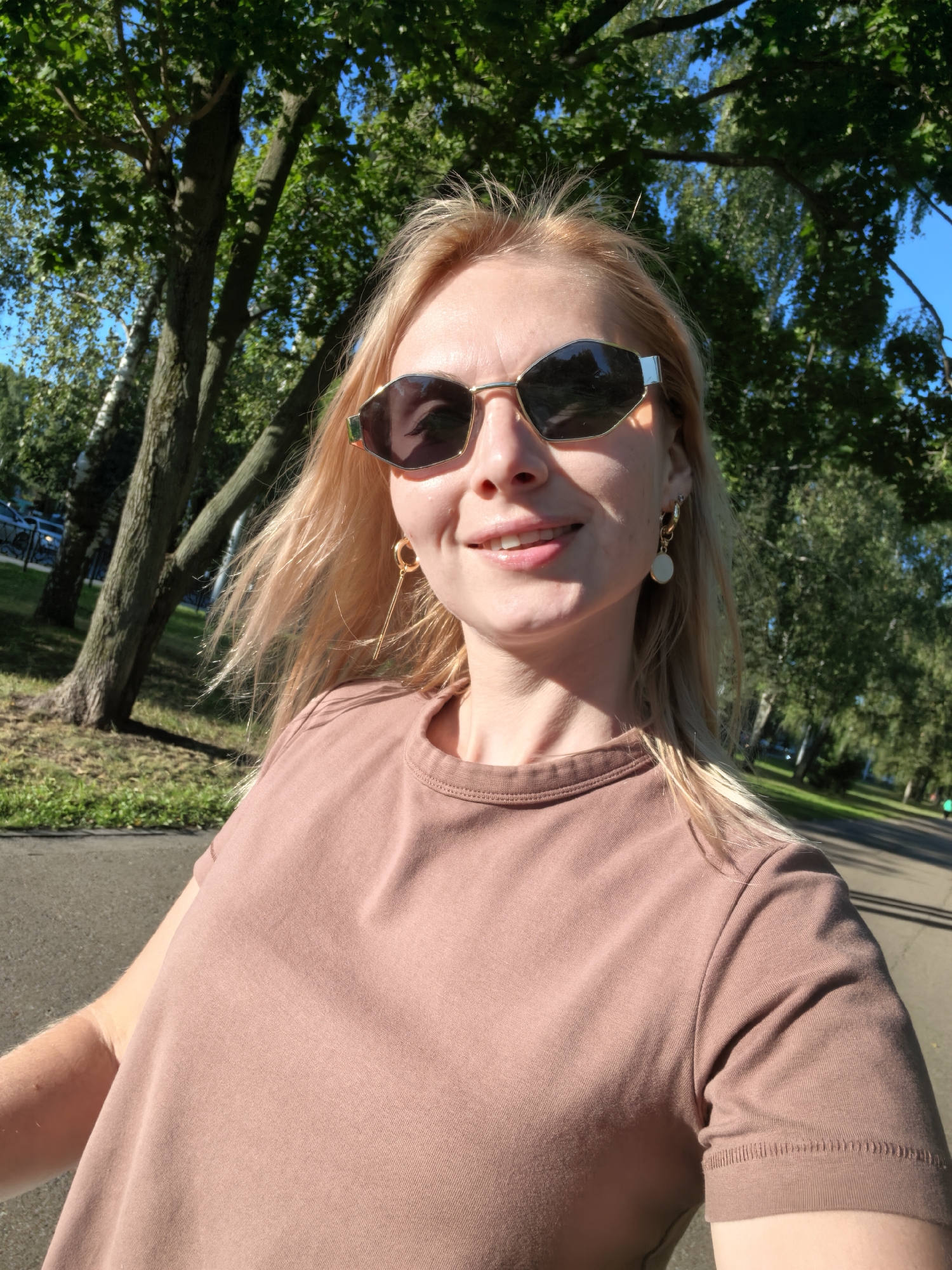 In Sterlitamak, Russia, a blonde woman with fair skin and blue eyes is captured in a selfie outdoors. She is wearing a dusty rose t-shirt and stylish sunglasses with geometric, angular frames. A delicate gold earring with a circular charm hangs from her left ear. Her hair, a light blonde, is windswept, suggesting a gentle breeze. She is smiling and looking directly at the camera. The background is dominated by lush green trees and a clear blue sky, indicative of a sunny day. The setting appears to be a park or a tree-lined pathway, possibly a recreational area within the city. Sunlight casts shadows and highlights, creating a warm and natural lighting effect. The focus is on the woman's face and upper body, with the environment serving as a softly blurred backdrop.