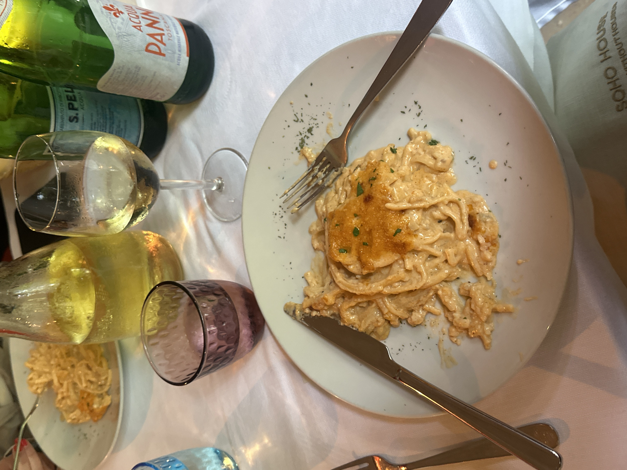 The image shows a table setting with a plate of pasta as the central subject. The pasta is topped with what appears to be a baked or grilled portion. A fork and knife are positioned on the plate, ready for use. The table is covered with a white tablecloth. Beverages are also present, including bottles of water, a glass of white wine, and an additional purple glass. The setting suggests a meal in a restaurant. A "Soho House" bag is partially visible, which is not in Venice, Italy. Therefore, no definitive determination of the location in the city of Venice, Italy, can be made.