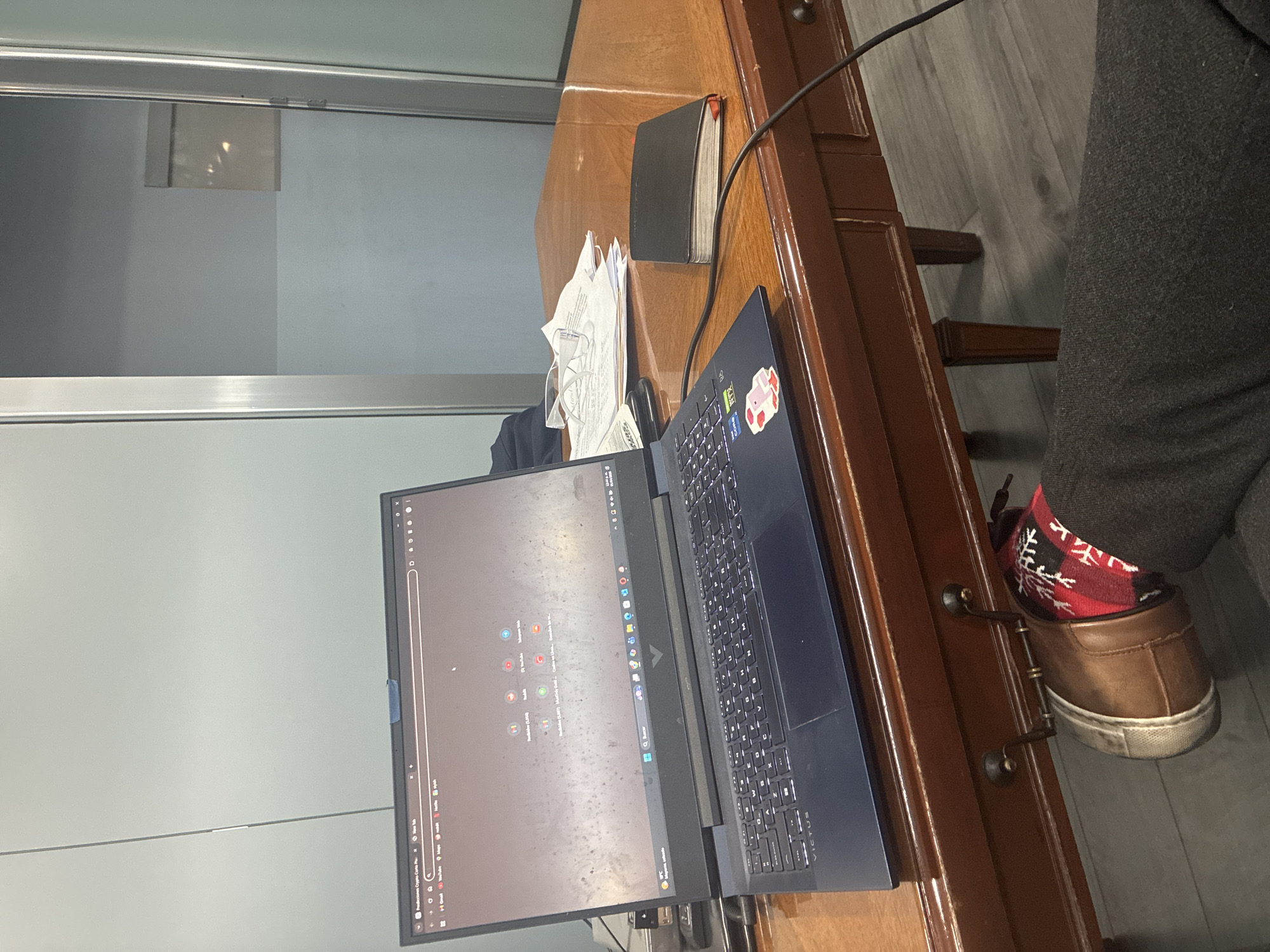 The image shows a desk in Cuautitlán Izcalli, Mexico. A laptop computer is open on the desk. A notebook and papers are next to it. A person is seated at the desk, wearing brown shoes and red and white socks. A window is visible in the background.