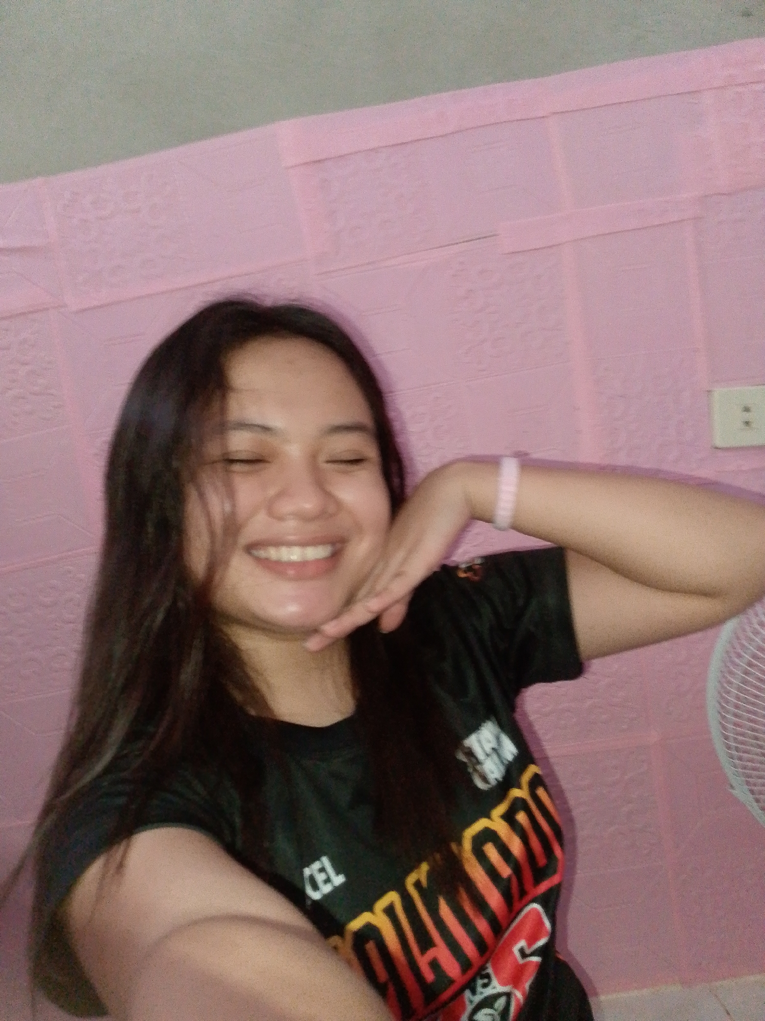 A young woman with long, dark hair is pictured from the chest up, smiling with her eyes closed. She is wearing a black t-shirt with red and yellow lettering. Her left hand is raised, resting on her chin. She is wearing a light pink bracelet on her left wrist. The background is a pink wall with a textured pattern. To the right of the woman, a portion of a white fan is visible. An electrical outlet with two sockets is mounted on the pink wall behind her right shoulder. The image is taken in an indoor setting with artificial lighting.