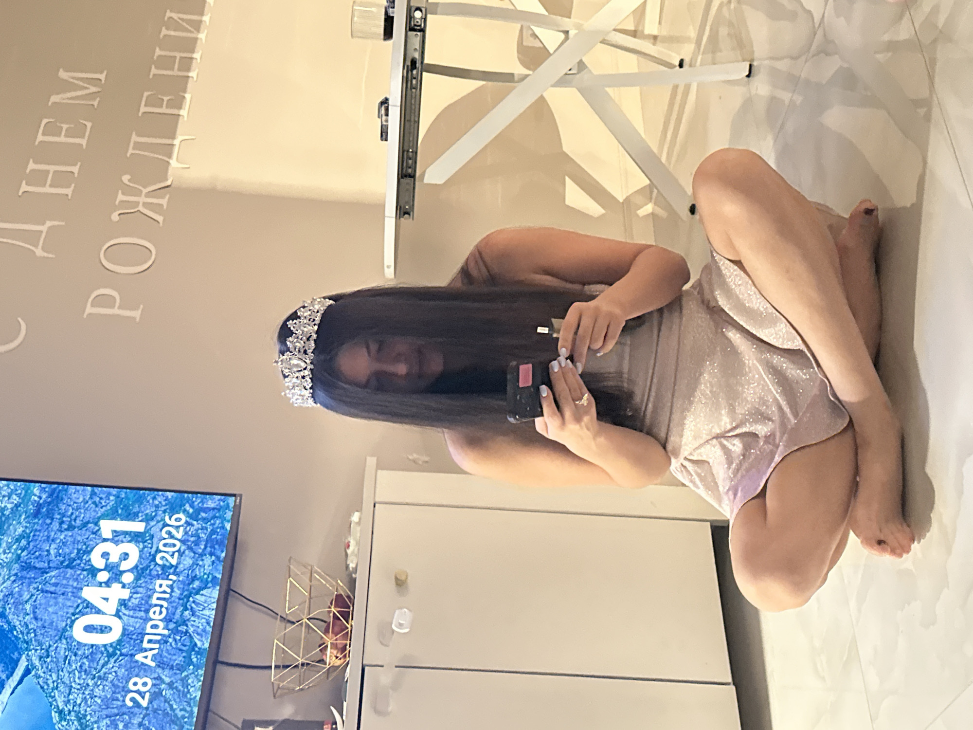 The image shows a young woman seated on a light-colored tiled floor in a modern indoor setting, likely an apartment in Moscow, Russia. She has long, dark hair and is wearing a sparkly tiara, along with a light-colored, possibly shimmery dress or top with shorts. In her hands, she holds a smartphone and a small makeup item, appearing to be applying makeup or checking her reflection. A ring is visible on her left hand's ring finger, and her bare feet have dark-painted toenails.
On the wall behind her, white dimensional letters spell out "С ДНЕМ РОЖДЕНИЯ," which translates to "Happy Birthday" in Russian, indicating a celebratory event. To the left, a television screen displays "04:31 28 Апреля, 2026" (04:31 April 28, 2026), showing the precise time and date against a blue, rocky landscape. The furniture includes a white desk or table with metal legs and a white cabinet. A gold geometric wire basket is visible near the TV. The woman's posture is relaxed, and her expression suggests focus or calm during what appears to be a birthday celebration.