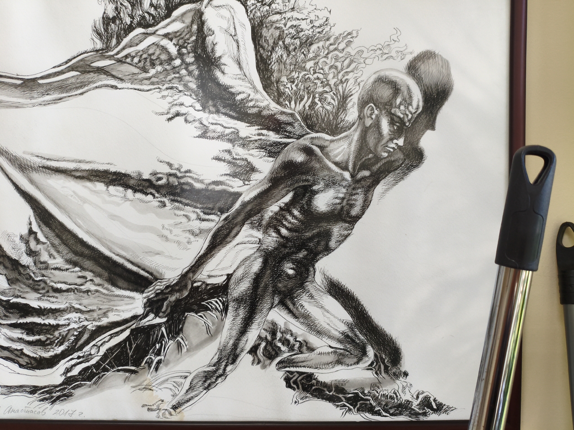 The image is a close-up, eye-level, indoor shot of a framed ink drawing. The drawing depicts a muscular, bald male figure with a wing-like appendage emerging from his back. The figure is rendered in a dynamic, contorted pose, with a sense of movement and struggle. The artistic style is highly detailed, utilizing a strong contrast between light and shadow created through hatching and cross-hatching techniques. The background of the drawing is largely white, suggesting an ethereal or abstract setting.
In the foreground, to the right of the drawing, parts of two cleaning implements are visible. One is a silver metal pole with a black plastic handle, and the other is a dark gray handle of a similar cleaning tool. The drawing is framed with a dark red border.
The image appears to be taken in an indoor setting, possibly a home or an art studio, as indicated by the presence of household items and the overall lighting. There is no direct indication of the specific location within Burgas, Bulgaria, however, the presence of a signature and date on the artwork, "Анастасов 2017 г.", suggests a Bulgarian artist.