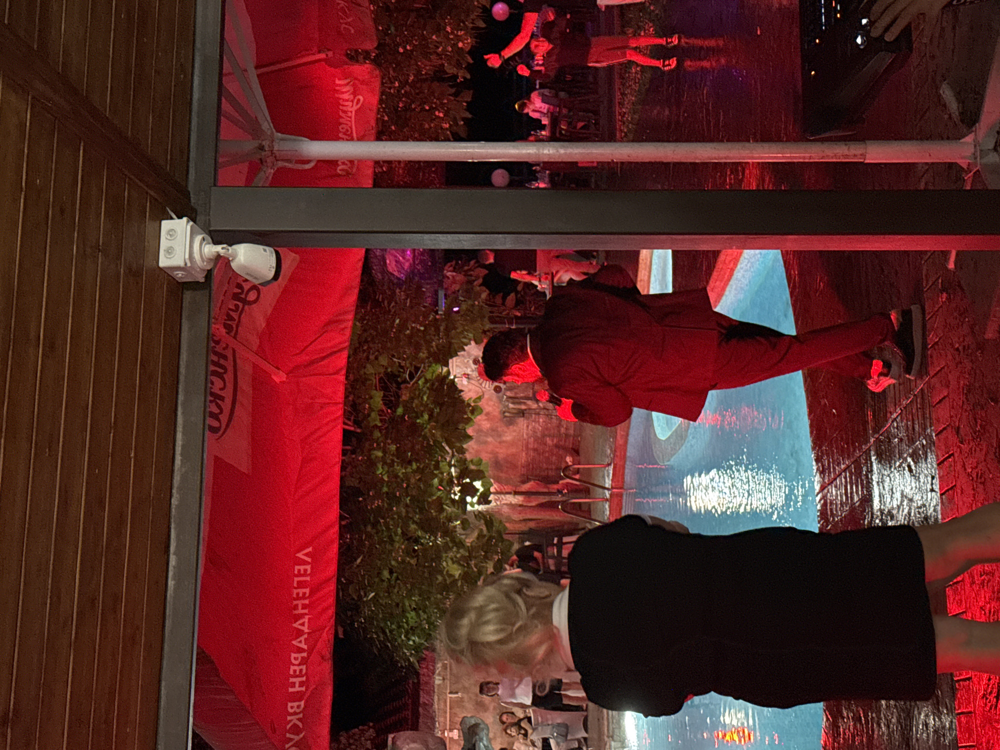 The image depicts an outdoor evening gathering, likely at a venue in Sredets, Bulgaria. The scene is illuminated by vibrant red lighting, casting a strong glow on the surroundings. In the foreground, a woman with blonde hair, wearing a black dress, stands with her back to the camera, facing a swimming pool. To her right, a man in a dark suit is speaking into a microphone, facing away from the camera and towards the pool area. Behind them, a group of people are visible in the background, seated at tables and standing near the pool. Umbrellas with red fabric are spread across the area, some displaying Cyrillic lettering that appears to say "VELEHNYA" and "BKAC". A security camera is mounted on the wooden ceiling above. The ground around the pool appears to be wet, possibly from recent rain or being sprayed. The overall atmosphere suggests a lively event with entertainment taking place.