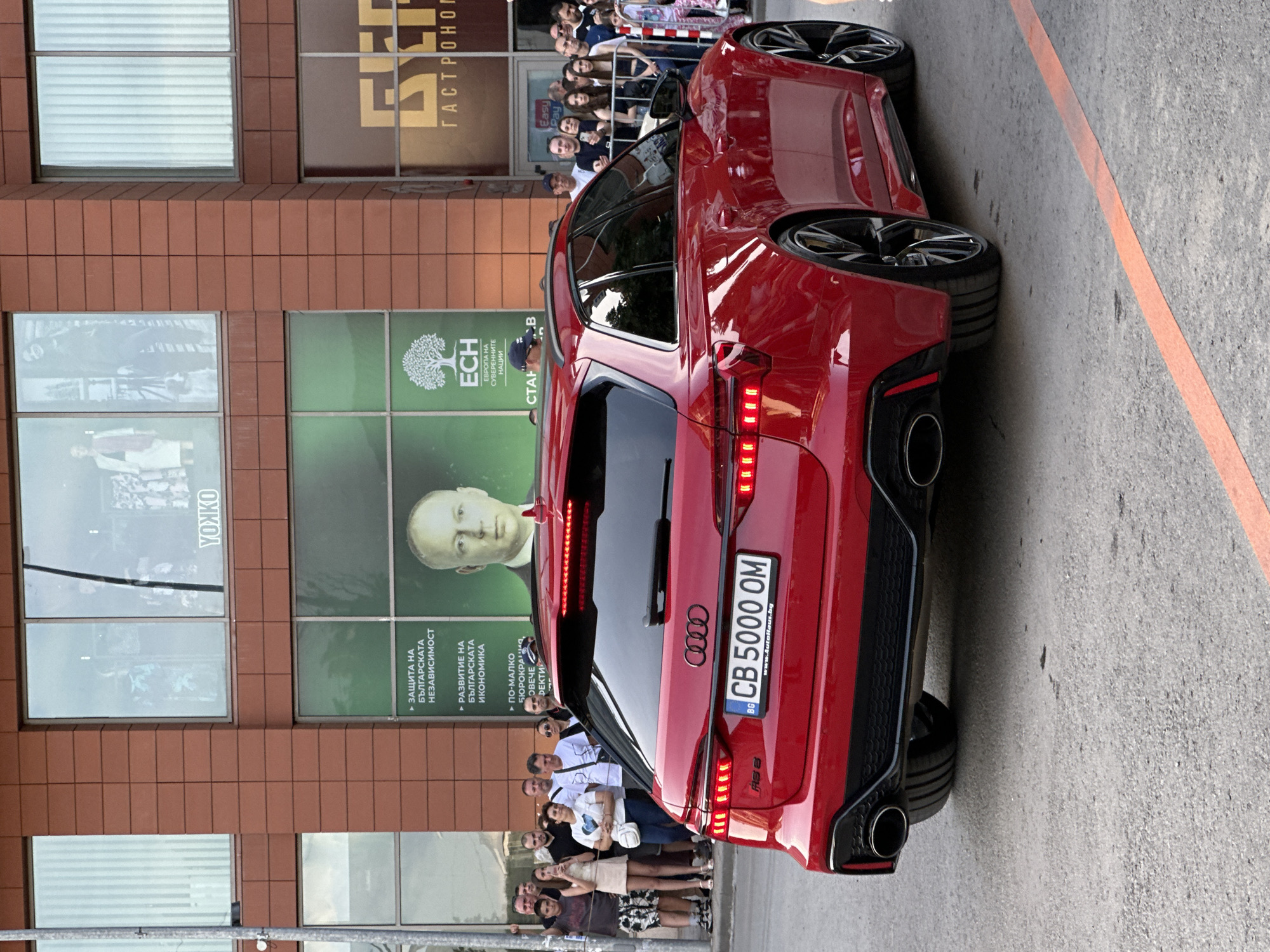 Here's a description of the scene:
The image shows a red Audi RS6 Avant parked on an asphalt surface with a faint orange line visible. A group of people are standing behind the car, partially visible, suggesting they might be looking at or admiring the vehicle.
Behind the people and the car, there is a multi-story building with a brick facade. Several windows or panels are visible on the building. One section has a large advertisement with a person's face (likely a politician or public figure) and a logo with a tree. Text in Bulgarian can also be seen, referencing "Protection of Bulgarian Independence" and other economic and political goals. Another window appears to display an advertisement. The text above is "ГАСТРОНОГ".
The license plate of the car reads "CB 5000 OM," indicating that the car is registered in Bulgaria, likely in the city of Sofia ("CB" prefix). There are other signs for "Easy Pay".
The location is determined to be in Veliko Tarnovo, Bulgaria, based on the presence of YONKO and other indicators within the images.