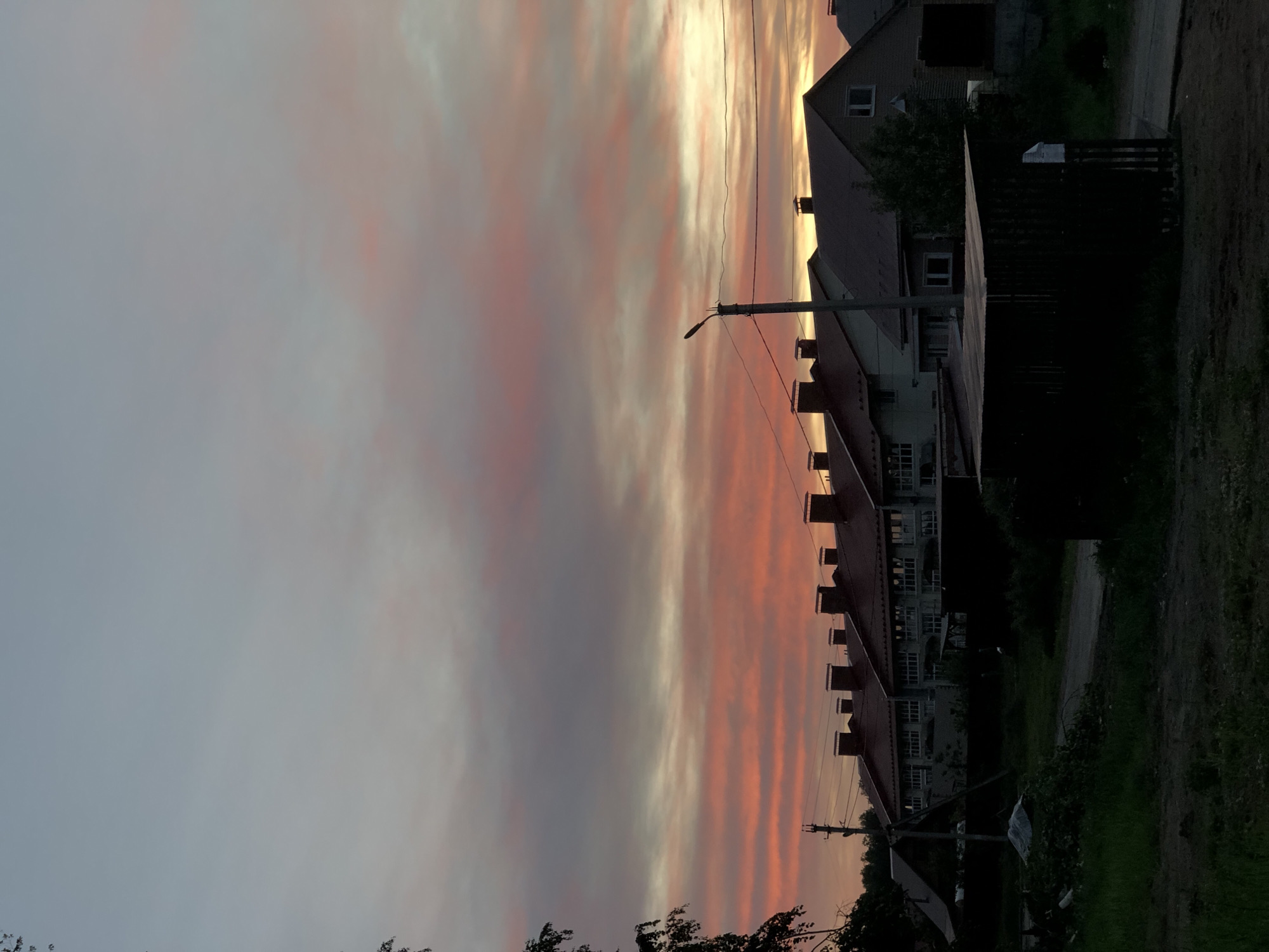 The image depicts a view in Noviy, Russia, at twilight or dawn, with buildings lining a street under a sky striped with grey, pink, and orange clouds. The houses have red roofs and white walls, with chimneys visible against the colorful sky. Utility poles and wires stretch across the scene, intersecting with the sky and rooftops. A dark fence and green foliage are seen on the side of the street.