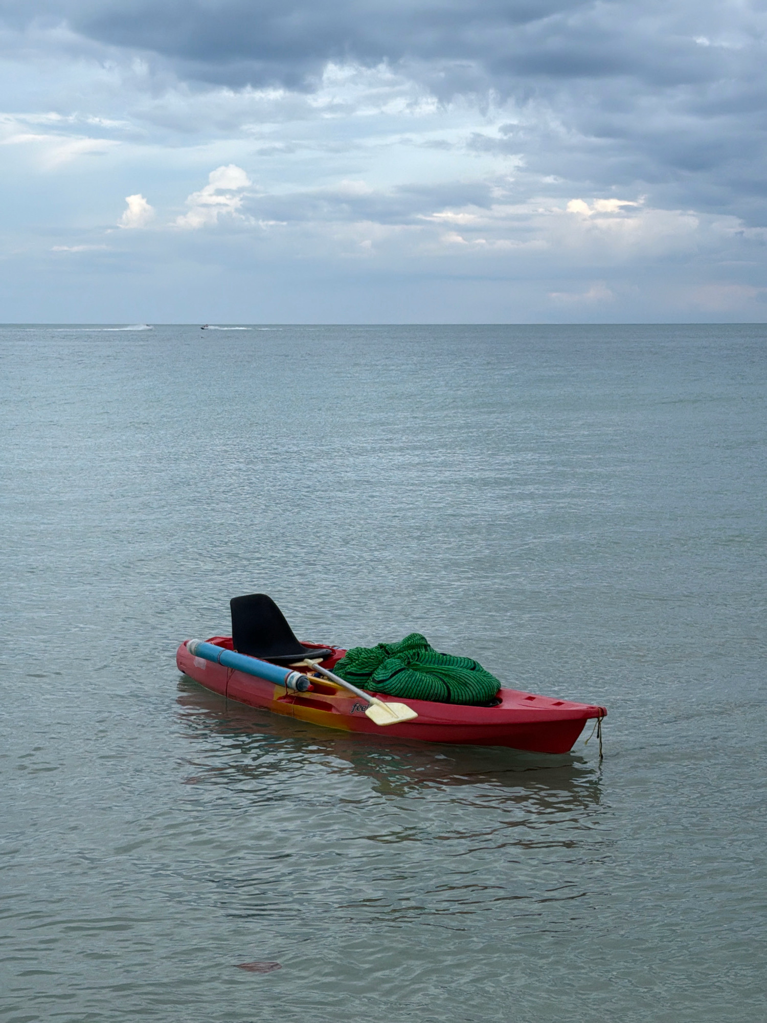 A red and yellow kayak with a black seat is adrift on a calm, gray sea under a cloudy sky. A green coiled rope and a paddle are placed inside the kayak. In the distance, two jet skis are visible on the water. The scene appears to be along the coast of Ko Samui District, Thailand, given the calm sea and tropical atmosphere.