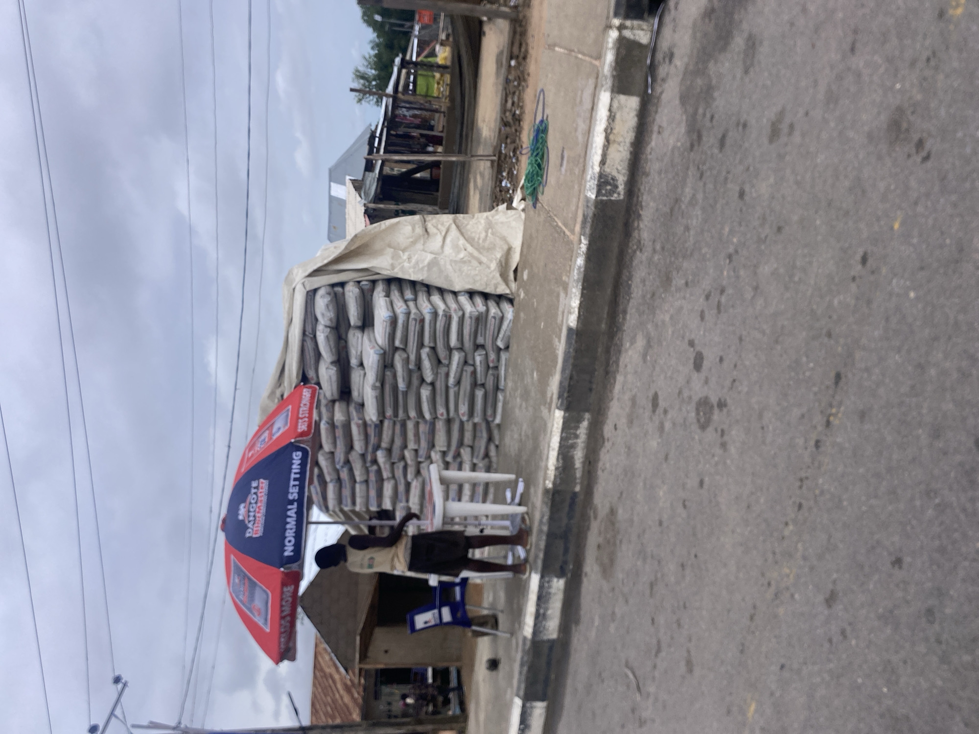 The image depicts an outdoor scene, likely a roadside market, in Jalingo, Nigeria. A large stack of cement bags, covered partially by a tan tarp, dominates the right side of the frame. A red and blue umbrella, advertising "DANGOTE BIOMASTER NORMAL SETTING SETS STRONGER," shades a small white table. A dark-skinned individual wearing a tan shirt and dark shorts stands behind the table, seemingly attending to the goods. To the left of the umbrella and table, a blue chair is visible next to a small structure. In the background, several more small buildings with corrugated metal roofs line the street, suggesting a commercial or residential area. The ground is paved with asphalt and a concrete curb. The sky is overcast.