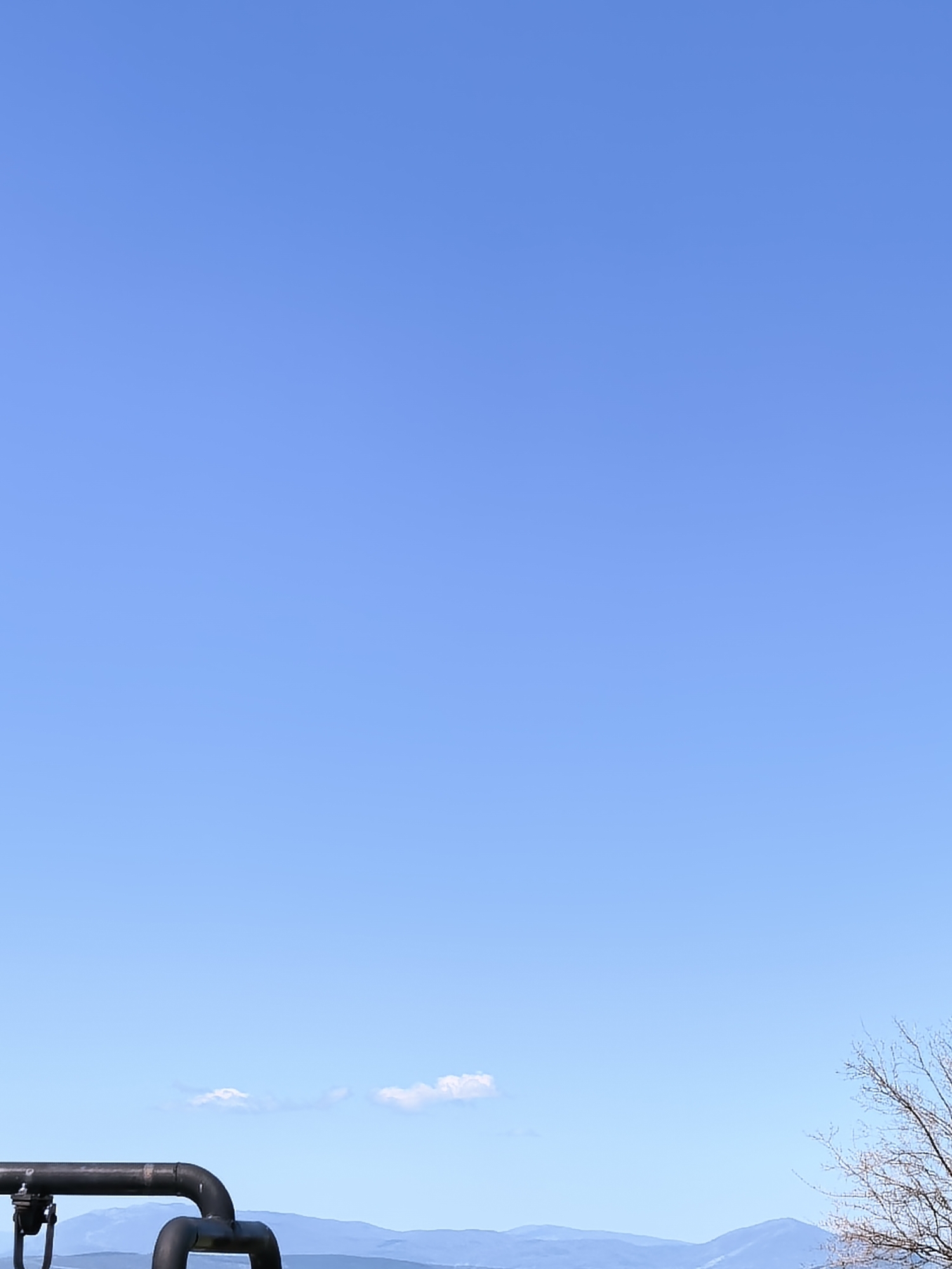 The image depicts a clear blue sky above a mountainous landscape. In the foreground, there is a black pipe system running horizontally and bending downwards. To the right of the pipes, a tree with bare branches is visible. A few small, white clouds are scattered above the mountains. The location is Unknown, Unknown.