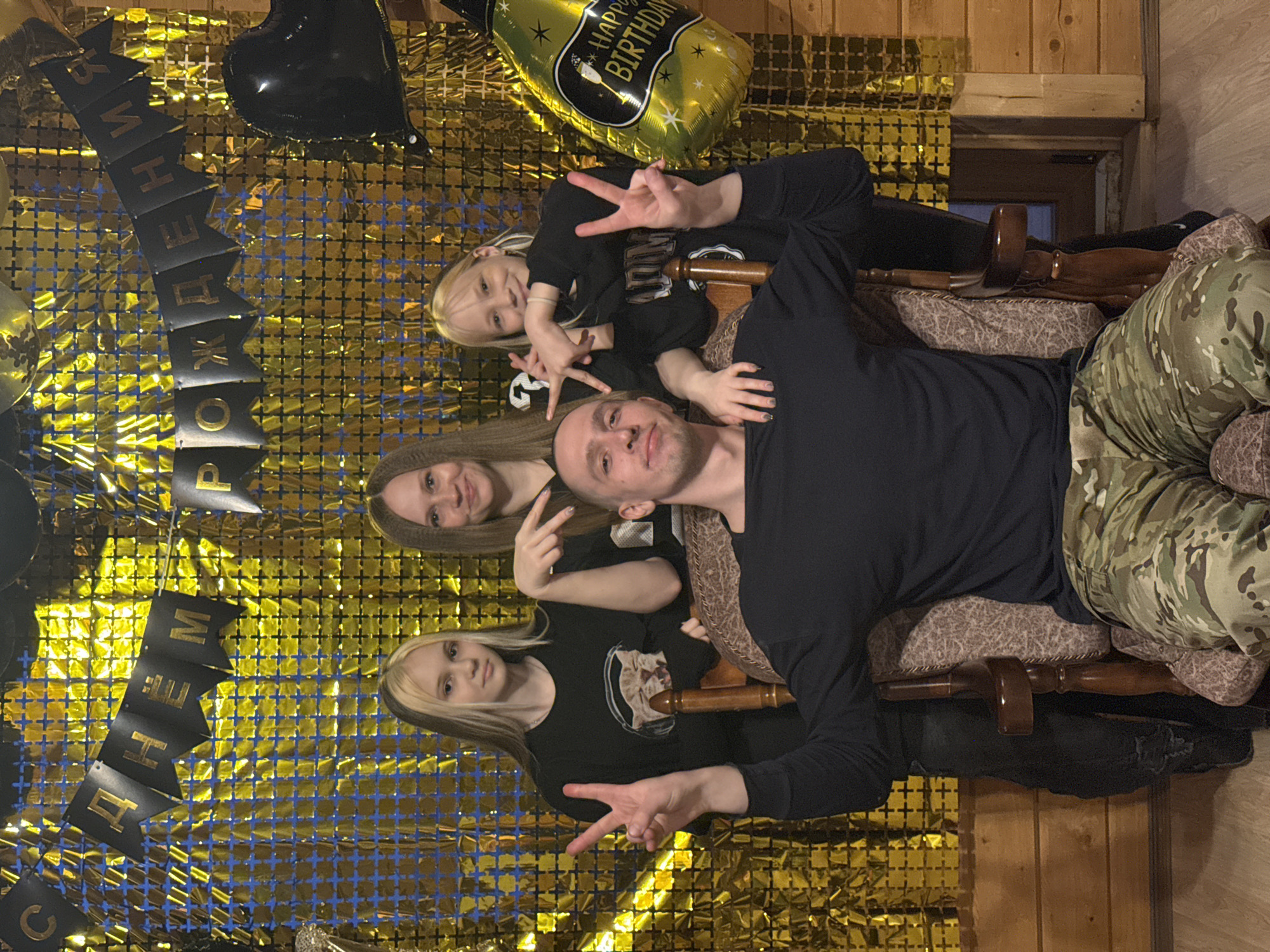 The image captures four individuals—a man, two women, and a young girl—posing for a photograph during a birthday celebration in what appears to be a casual indoor setting, likely a home in Chaikovsky, Russia. The group is seated or reclining on two patterned armchairs or sofa sections, with a rustic wood-paneled wall visible behind them on the right.
The backdrop for the celebration is a shimmering gold tinsel curtain layered over a blue grid pattern. Decorations include a black heart-shaped balloon and a gold champagne bottle-shaped balloon, which clearly displays "Happy Birthday" text. A prominent black banner with gold lettering hangs across the golden backdrop, reading "С ДНЁМ РОЖДЕНИЯ," which is Russian for "Happy Birthday."
The man, with a shaved head, is in the center-right, wearing a dark long-sleeved shirt and camouflage pants. The young girl, with blonde hair, is nestled between the man and one of the women, wearing a black shirt and making a peace sign with both hands. The woman to the left of the man has long brown hair and is smiling, also making a peace sign. The other woman, with long blonde hair, is on the far left, wearing a black shirt with a cat graphic and making a peace sign. All individuals appear happy and relaxed, with most showing smiles or offering peace gestures to the camera. The indoor lighting suggests the event is taking place in the evening or under artificial light.