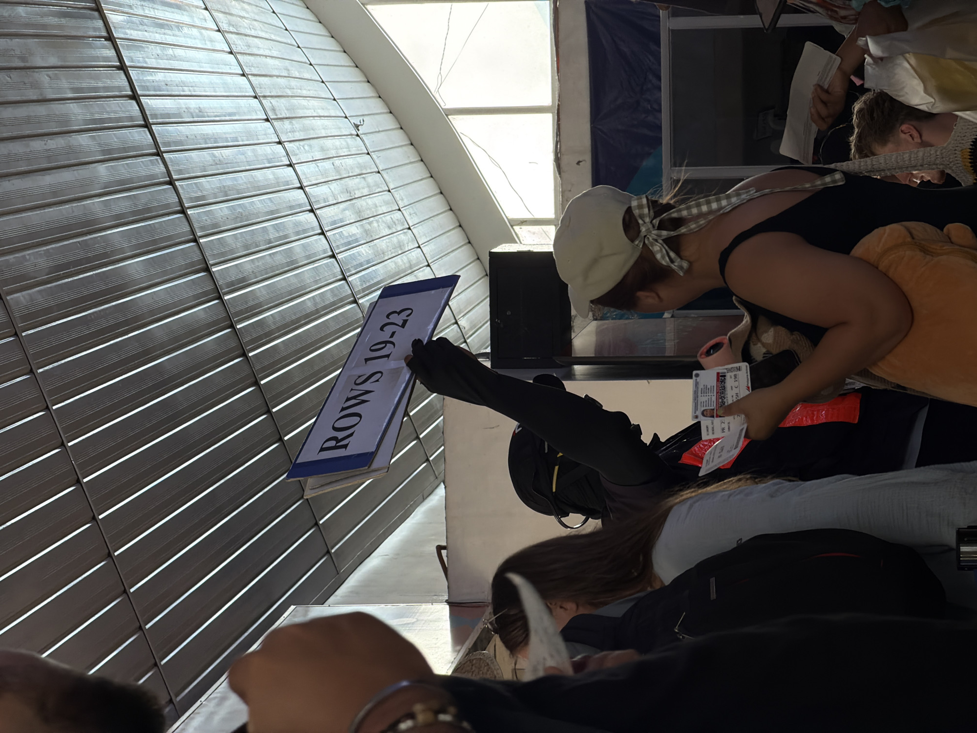 The image shows an indoor scene, likely a seating area or waiting room, potentially in Del Carmen, Philippines. People are present, with a woman wearing a cap and black tank top. Another individual is partially visible, wearing black clothing and potentially a backpack. Someone is holding a sign displaying "ROWS 19-23". The setting features what appears to be a slatted wall or ceiling structure. The overall scene suggests a process of checking or directing people to specific rows, possibly at an event or transportation hub.