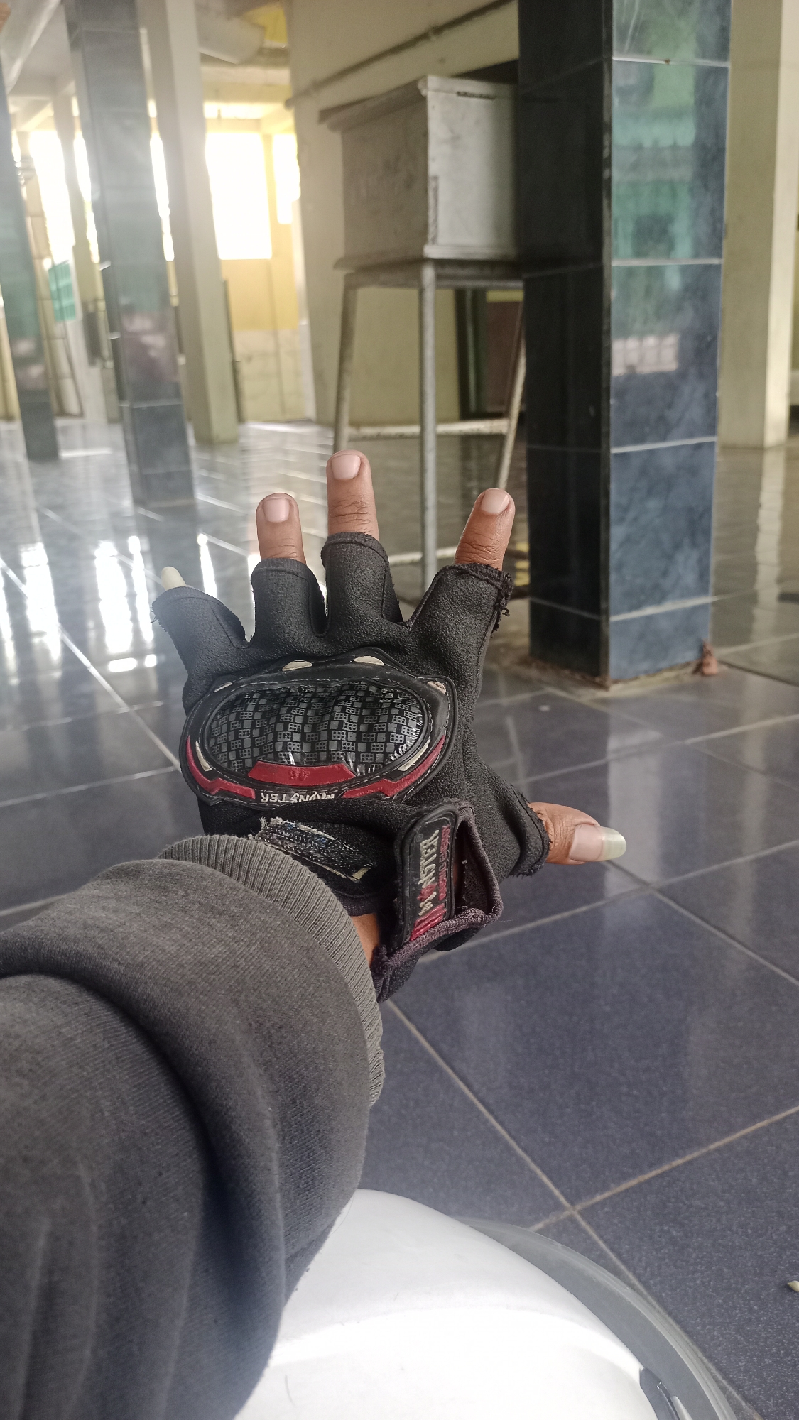 A person's left hand wearing a black fingerless glove with red and silver accents is prominently displayed in the foreground. The glove has a protective shell on the back of the hand and features a checkered pattern. The hand is held up, with the index, middle, and ring fingers extended, while the thumb and pinky are slightly bent. The person is wearing a grey long-sleeved shirt or sweatshirt. The background suggests an indoor or covered outdoor space with tiled flooring and large pillars. Sunlight streams in from the left, creating reflections on the floor. In the distance, a metal structure resembling a large bin or container on a stand is visible. The scene appears to be within a public or communal area, possibly a mosque or a similar building, given the architecture. The location is Karyamandala, Indonesia.
