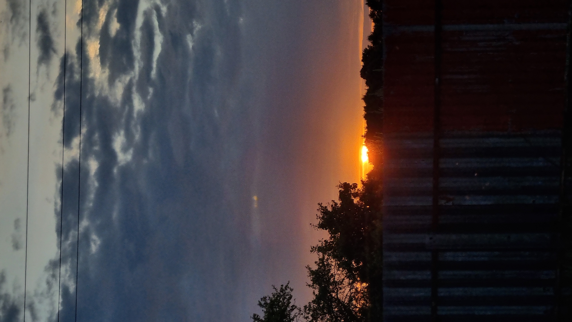 The image captures a sunset scene in Vysokoe, Russia, viewed over a corrugated metal fence. The sky is filled with dramatic clouds, transitioning from dark, heavy blues at the top to lighter grays and pinks closer to the horizon. The sun, a bright orange orb, is partially visible just above a silhouetted treeline, casting a warm glow that illuminates the lower sky. The fence in the foreground is dark and textured, with vertical ridges and hints of reddish-brown rust. Trees are visible in silhouette against the sunset, with some branches extending into the upper sky. Power lines are strung horizontally across the top of the frame.